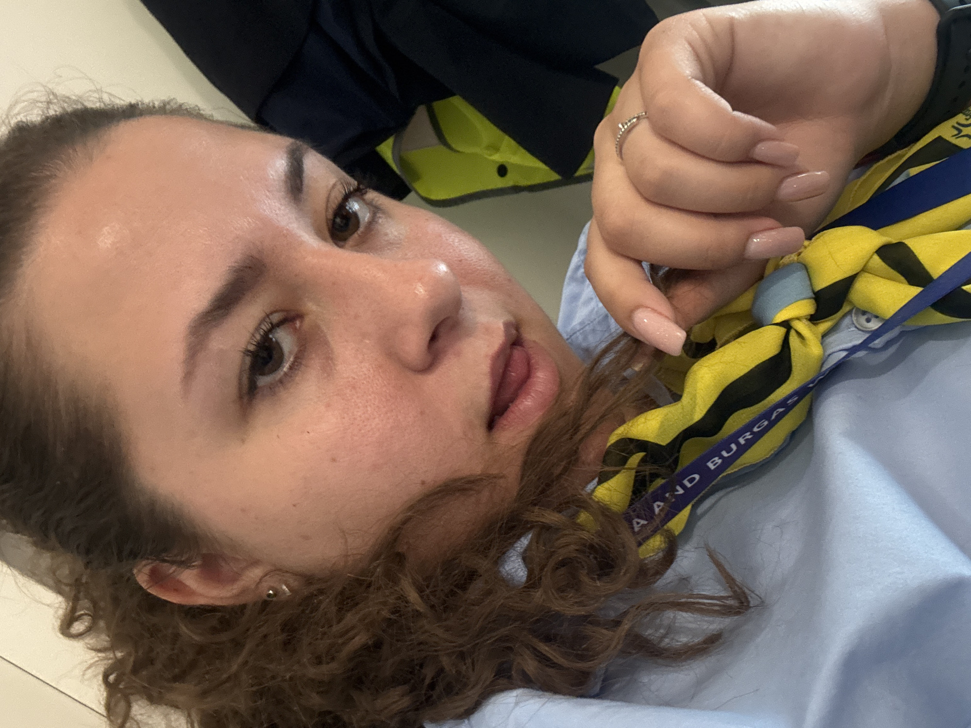 A woman with curly brown hair, wearing a light blue collared shirt and a yellow and black striped tie with blue lettering, is seen from the chest up. She has dark brown eyes and her tongue is slightly protruding from her lips. A silver ring with a small jewel is on her right ring finger. The background shows dark navy and bright yellow fabric, possibly clothing or uniforms. The image appears to be taken indoors, with soft lighting. The woman's posture suggests a casual selfie.