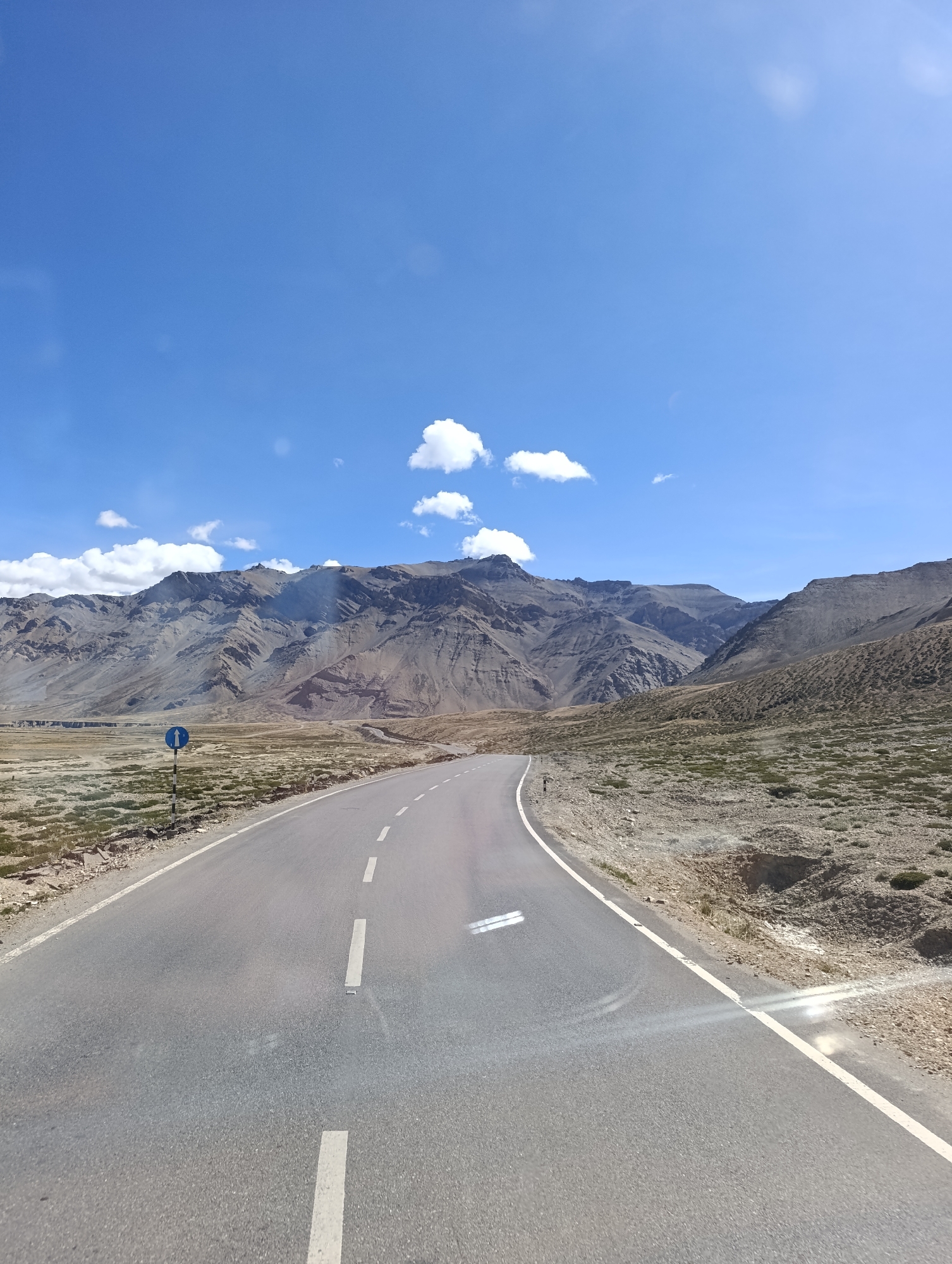 The image depicts a winding asphalt road through a mountainous, arid landscape under a bright blue sky with scattered white clouds. A blue and white road sign with an arrow indicating a turn is visible on the left side of the road. The mountains in the background are rugged and appear to be made of rock and scree. The vegetation is sparse, consisting of low-lying shrubs and dry grasses. The road has white dashed lines indicating lanes. The scene appears to be in a remote, high-altitude region.
