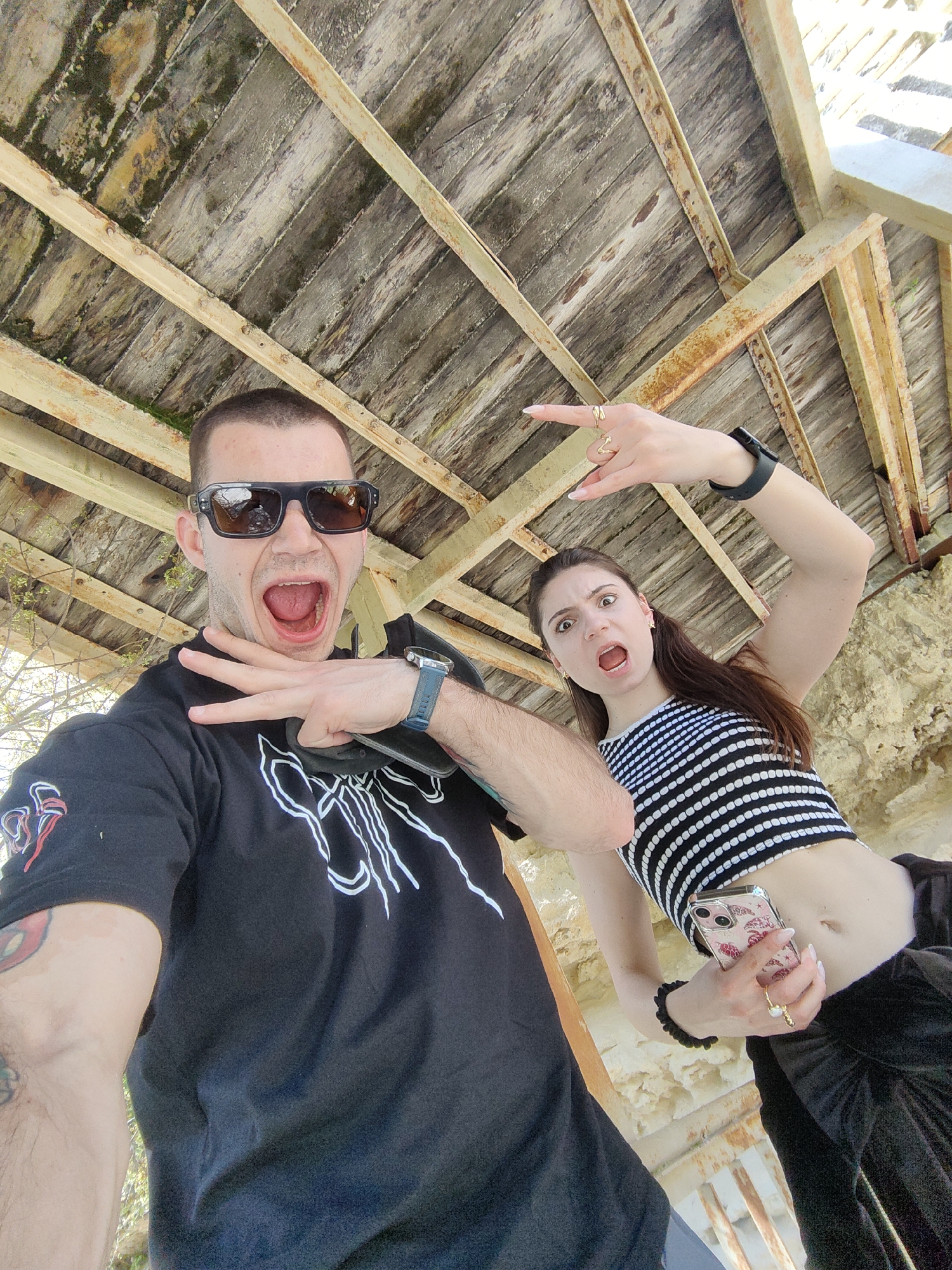 A selfie-style, wide-angle shot, possibly taken with a smartphone, captures two individuals under a dilapidated, wooden structure. The wooden planks of the ceiling are old, weathered, and in a state of disrepair, supported by rusted metal beams.
The man on the left wears sunglasses and a black t-shirt with a white design. He has a watch on his wrist and tattoos on his upper arm. He is facing the camera with his mouth wide open, making a hand gesture. The woman on the right is wearing a black and white striped top and dark pants. She has long brown hair and is also facing the camera with her mouth open, holding a smartphone with a clear case adorned with butterflies. She is making a "rock on" hand gesture with her right hand and has a watch on her left wrist.
The background appears to be an outdoor setting, with hints of foliage visible in the periphery. The photo was probably taken under a wooden structure. The precise location within Varna, Bulgaria, is impossible to determine without additional context.