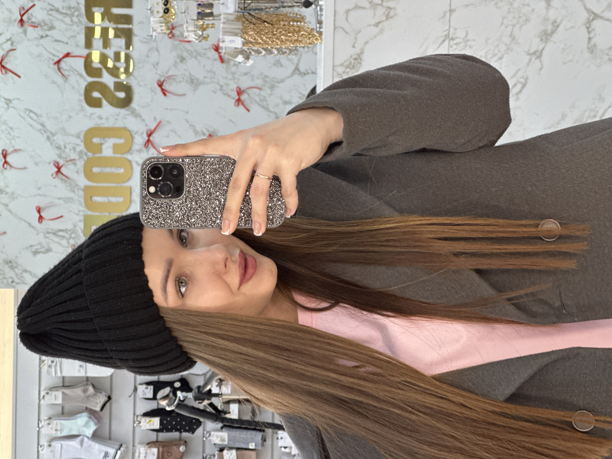 A young female, with long straight brown hair and light eyes, is depicted in an indoor setting, taking a selfie with a smartphone held in her left hand. She wears a black ribbed knit beanie, a pink t-shirt, and a dark grey open coat. Her left ring finger displays a slender ring, and her nails are manicured with a French tip style. The smartphone, an iPhone model with a three-camera system, is encased in a glittery dark grey cover. The background consists of a white wall with a subtle marble pattern, adorned with multiple small red bows and gold mirrored letters that partially spell "DRESS COD" (reversed). To the left of the subject, a retail display rack holds various packaged socks or small textile items. To the right, a stand showcases multiple necklaces, primarily gold-colored, and earrings. The scene is located in the city of Chaikovsky, Russia.