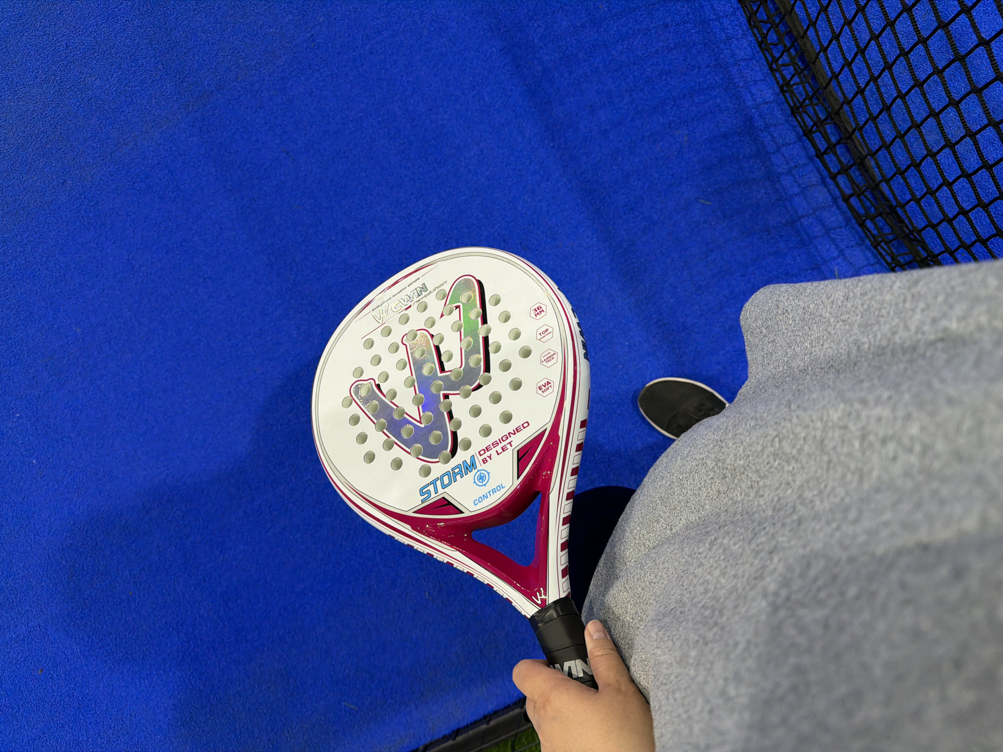 A top-down shot shows a person holding a white paddle tennis racket with pink accents. The racket is branded "VS WIN" and "STORM DESIGNED BY LET." The person is wearing grey clothing and a black shoe is visible. The background is a blue surface, possibly a tennis court, and netting. It is not possible to definitively confirm the location is Chaikovsky, Russia from the image alone.