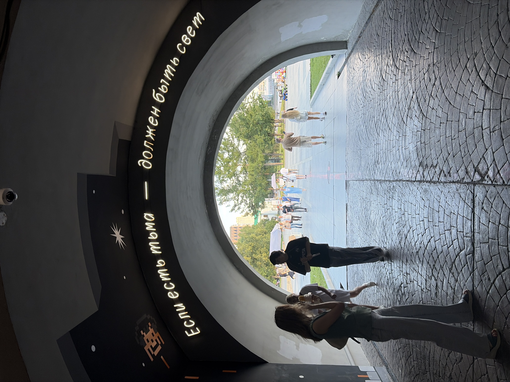 An outdoor, wide-angle shot captures a view through a large, curved archway, revealing a sunlit plaza beyond. The interior of the archway is dark and features a neon sign in Russian that reads "Если есть тьма — должен быть свет" (If there is darkness, there must be light). The ceiling of the archway is decorated with subtle celestial elements, including a pixelated spaceship graphic and a starburst.
In the foreground, inside the archway, a woman stands facing away from the camera, wearing a green top and light-colored pants. To her right, partially obscured, stands a man in a dark t-shirt and patterned pants, holding a microphone. Further back, a woman in white walks away from the camera.
The plaza outside the archway is paved with light-colored stone and features trees, buildings in the distance, and several people walking. Some individuals are holding ice cream cones. A white tent is visible in the middle ground. The lighting suggests it is daytime. The image appears to have been taken at the "Vysota 96" (Height 96) observation deck in Yekaterinburg, Russia, which is known for its panoramic views and artistic installations, including the archway with the neon inscription.