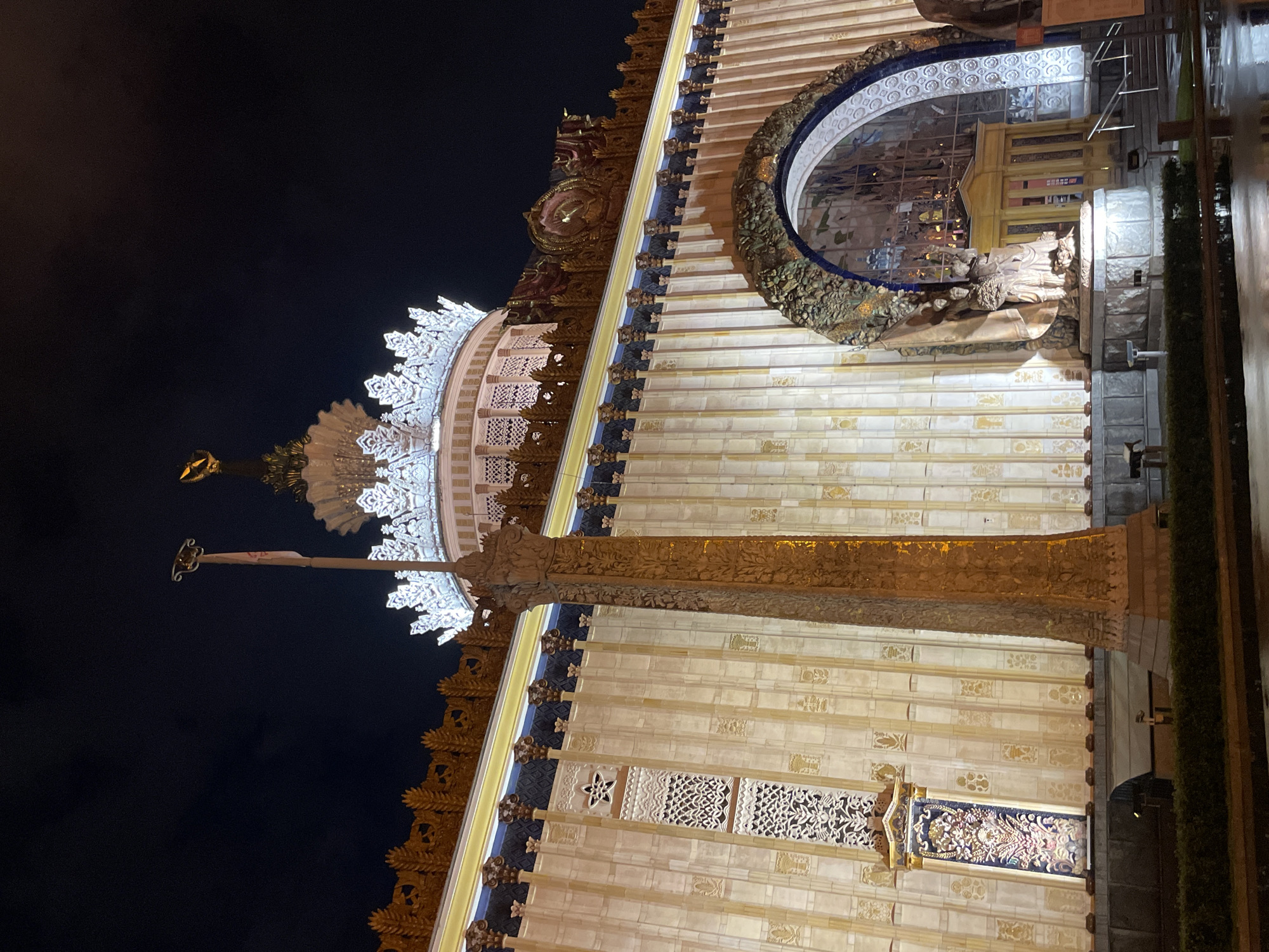 This is a nighttime shot of the exterior of Pavilion No. 1 “Central” at the Exhibition of Achievements of National Economy (VDNKh) in Moscow, Russia. The building is illuminated, showcasing its elaborate facade with white and gold ornamentation. At the top, there is a white, ornate crown-like structure with a golden statue on a pole extending from it. Below, the building's exterior features rows of horizontal panels, decorative windows, and a large, circular window with a sculpture visible inside. The right side of the frame shows a hint of greenery and a modern walkway or ramp. The sky in the upper left corner is dark and clear.
