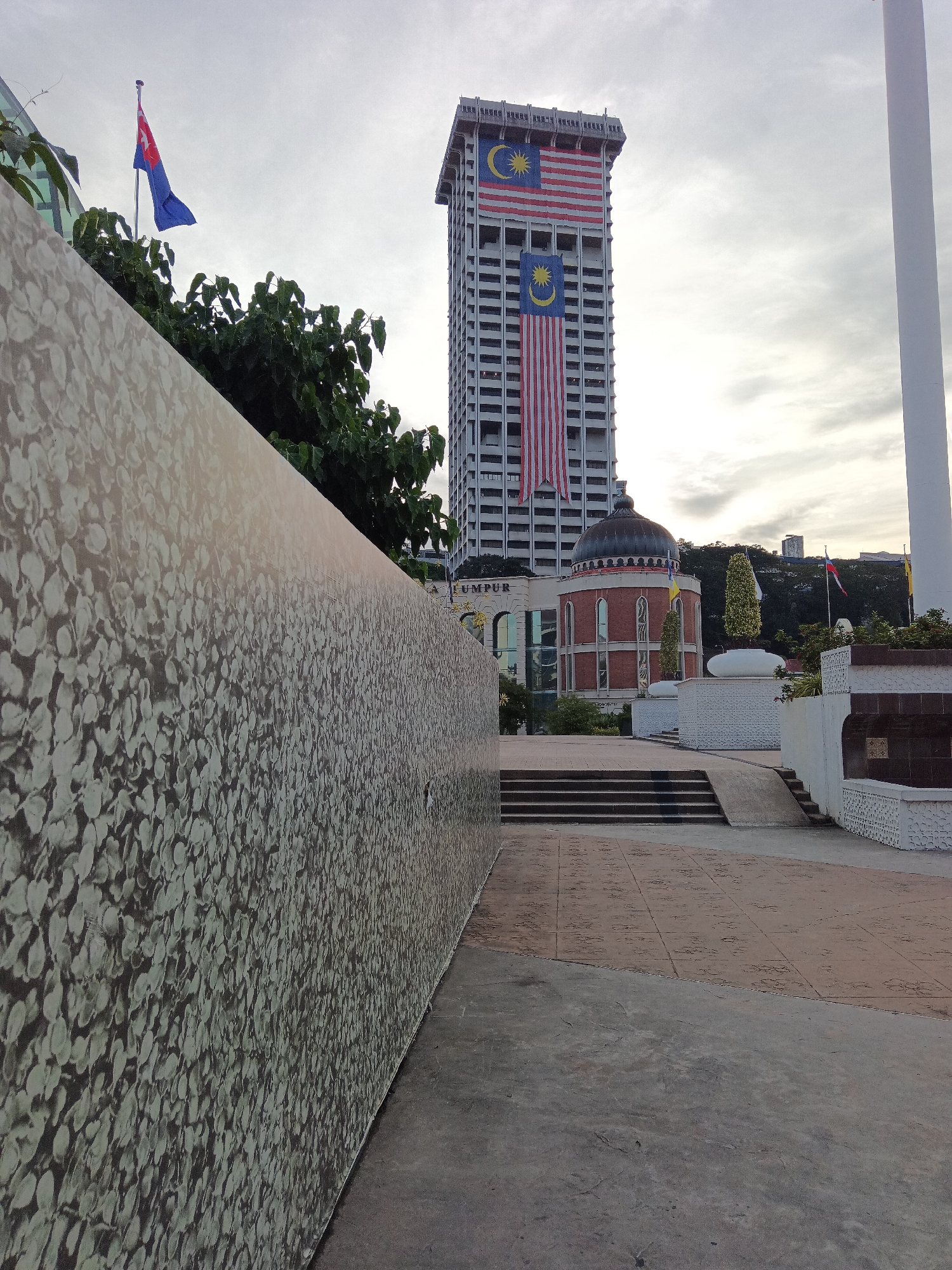 The image depicts a view of the Merdeka Square area in Kuala Lumpur, Malaysia. In the background, the iconic Sultan Abdul Samad Building stands tall, adorned with two large Malaysian flags. To the left of the building, a smaller flag, possibly a state flag, flutters on a pole. A large, patterned wall occupies the foreground, extending diagonally across the frame. The ground is paved with concrete and patterned tiles, with a set of stairs leading up towards the building. The sky is overcast, suggesting a cloudy day. The overall scene is an outdoor urban landscape, featuring architectural landmarks and patriotic symbols.