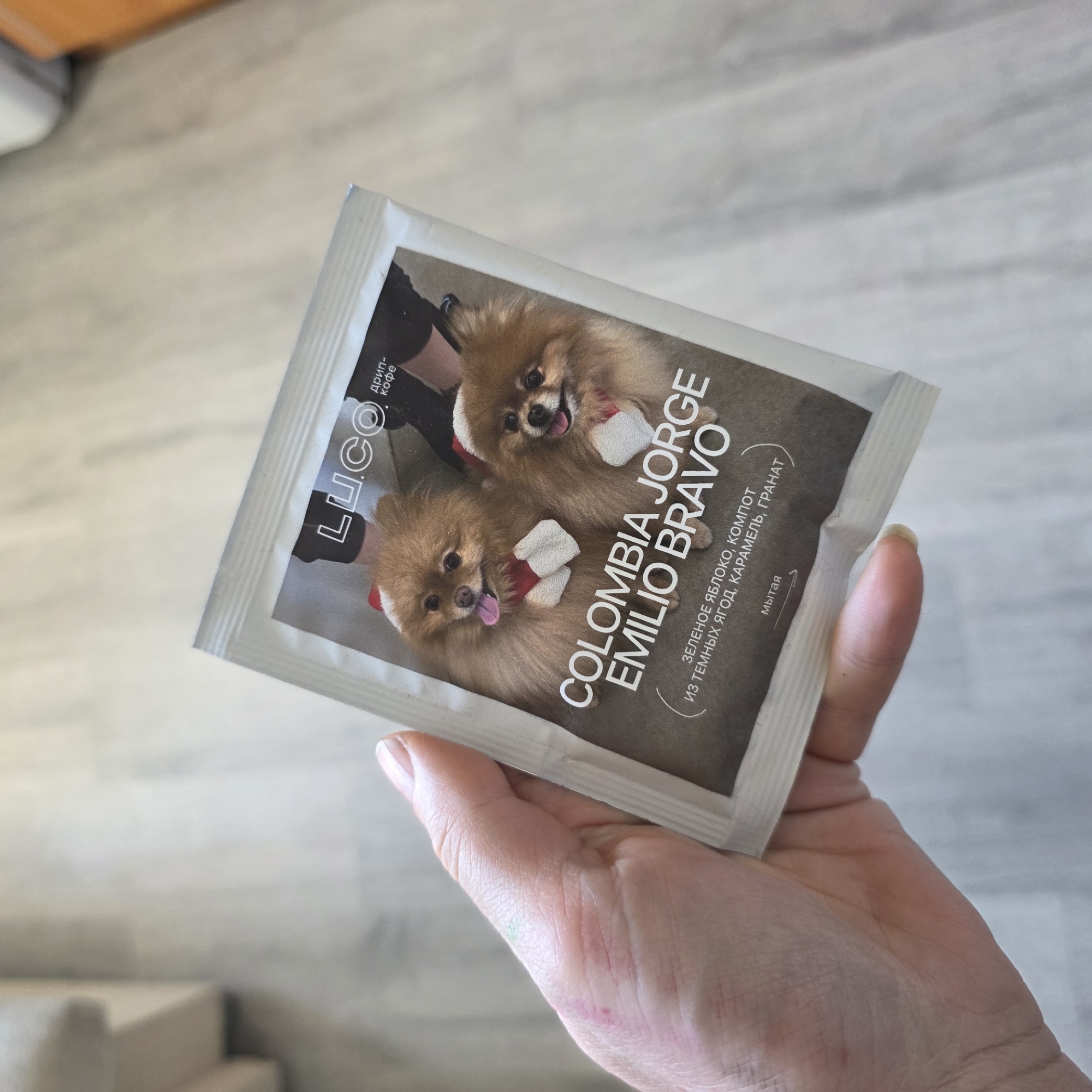 A hand is holding a small, white package with a picture of two dogs on it. The package has the words "LU.CO" and "COLOMBIA JORGE EMILIO BRAVO" printed on it in Russian. The text describes the coffee as having notes of green apple, dark berry compote, caramel, and pomegranate. The package is being held indoors, possibly in a residential setting. The location is the city of Saint Petersburg, Russia.