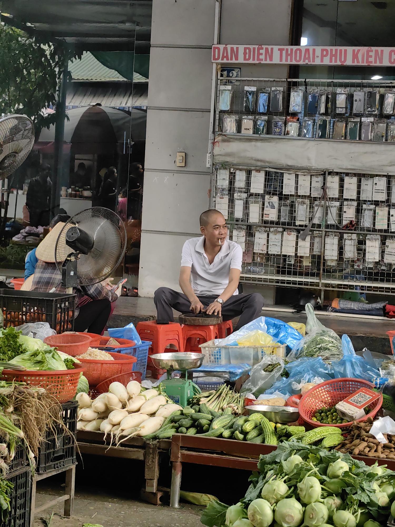 The image captures a bustling street scene in Ha Long Ward, Vietnam. In the foreground, a vendor sits on a red plastic stool amidst a vibrant display of fresh produce. The assortment includes kohlrabi, cucumbers, radishes, and other vegetables neatly arranged on wooden platforms. The vendor, a man with a shaved head, is dressed in a white polo shirt and dark trousers. He holds a cigarette in his mouth. Behind him is a rack filled with mobile phone accessories. To his left is another vendor wearing a conical hat and seated behind a large electric fan. The backdrop suggests a commercial area.