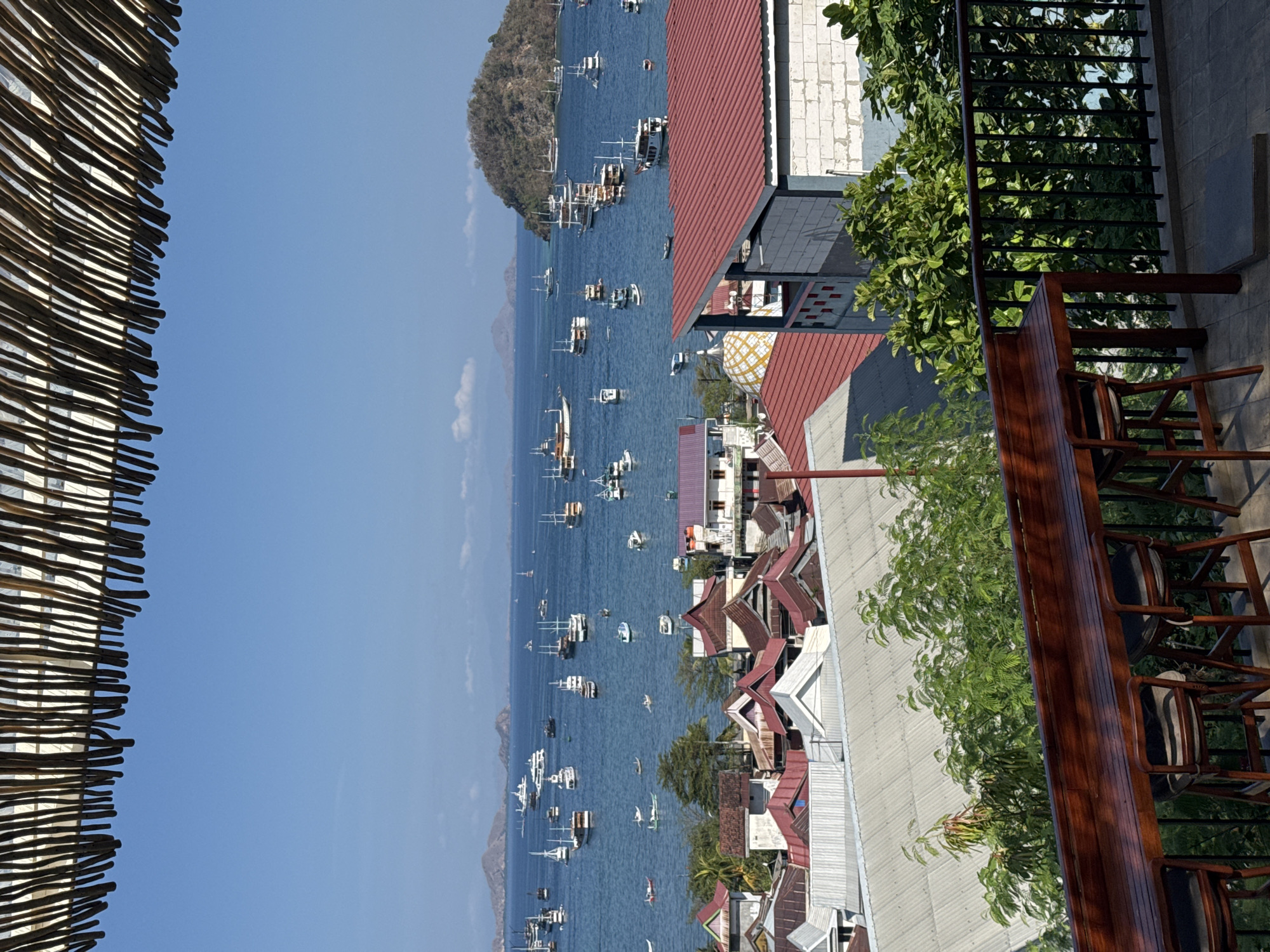 The image shows a scenic view of Labuan Bajo, Indonesia, from an elevated vantage point, possibly a restaurant or balcony. A wooden bar with several stools is in the foreground, overlooking a bay dotted with numerous boats and ships. The bay is calm and blue under a clear sky. In the middle ground, there is a coastal town with buildings featuring red and grey roofs. Beyond the town, a lush green hill rises from the sea. The overall scene is tranquil and picturesque, showcasing the natural beauty and maritime activity of Labuan Bajo.