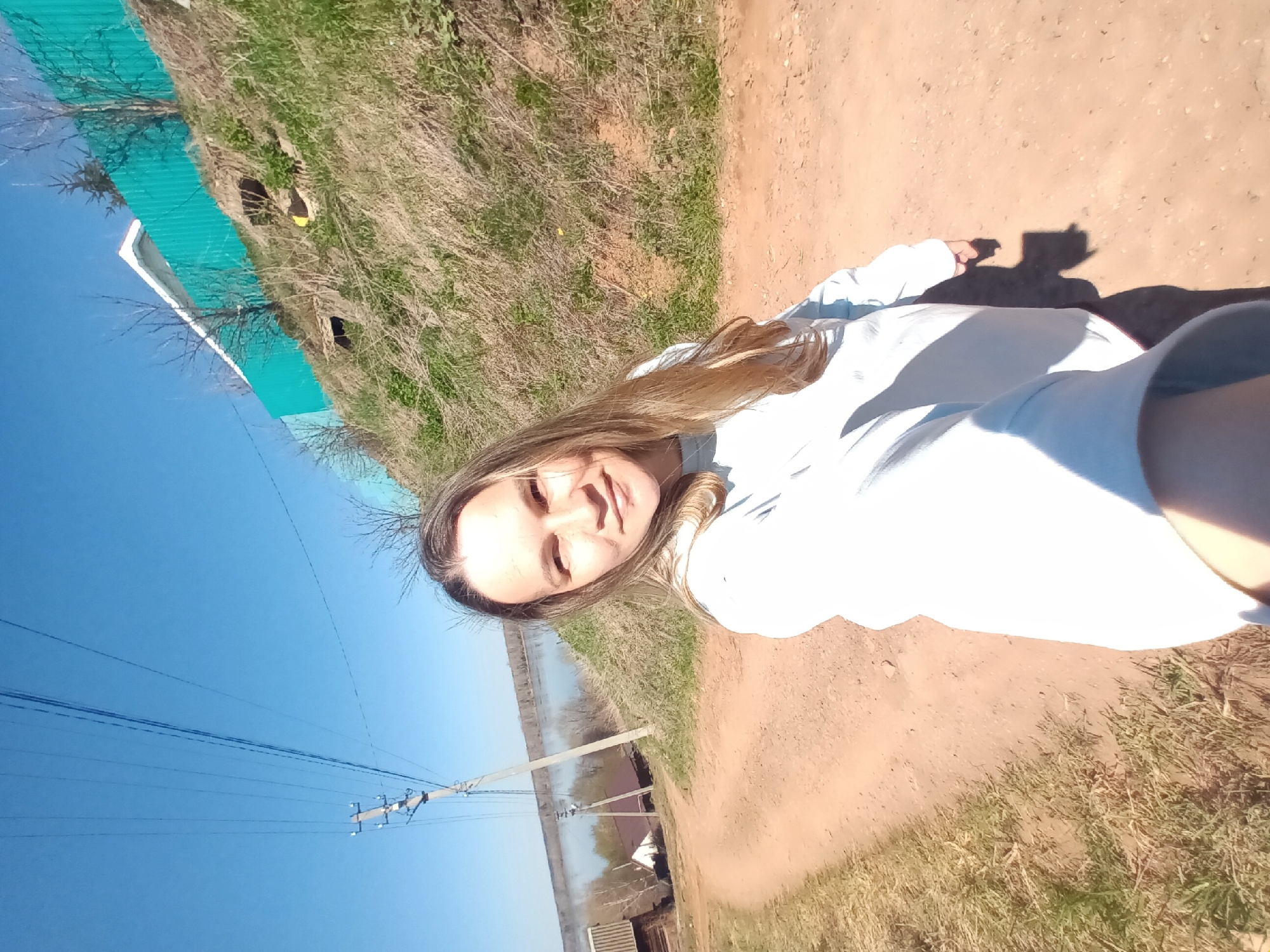 A young woman is photographed outdoors in what appears to be a rural area of Ольховка, Russia. She is standing on a dirt road, smiling at the camera, wearing a light-colored sweatshirt. The road is bordered by grassy areas with dry vegetation. In the background, there is a blue sky with power lines and poles, along with glimpses of buildings and possibly a body of water. The lighting indicates it is daytime.