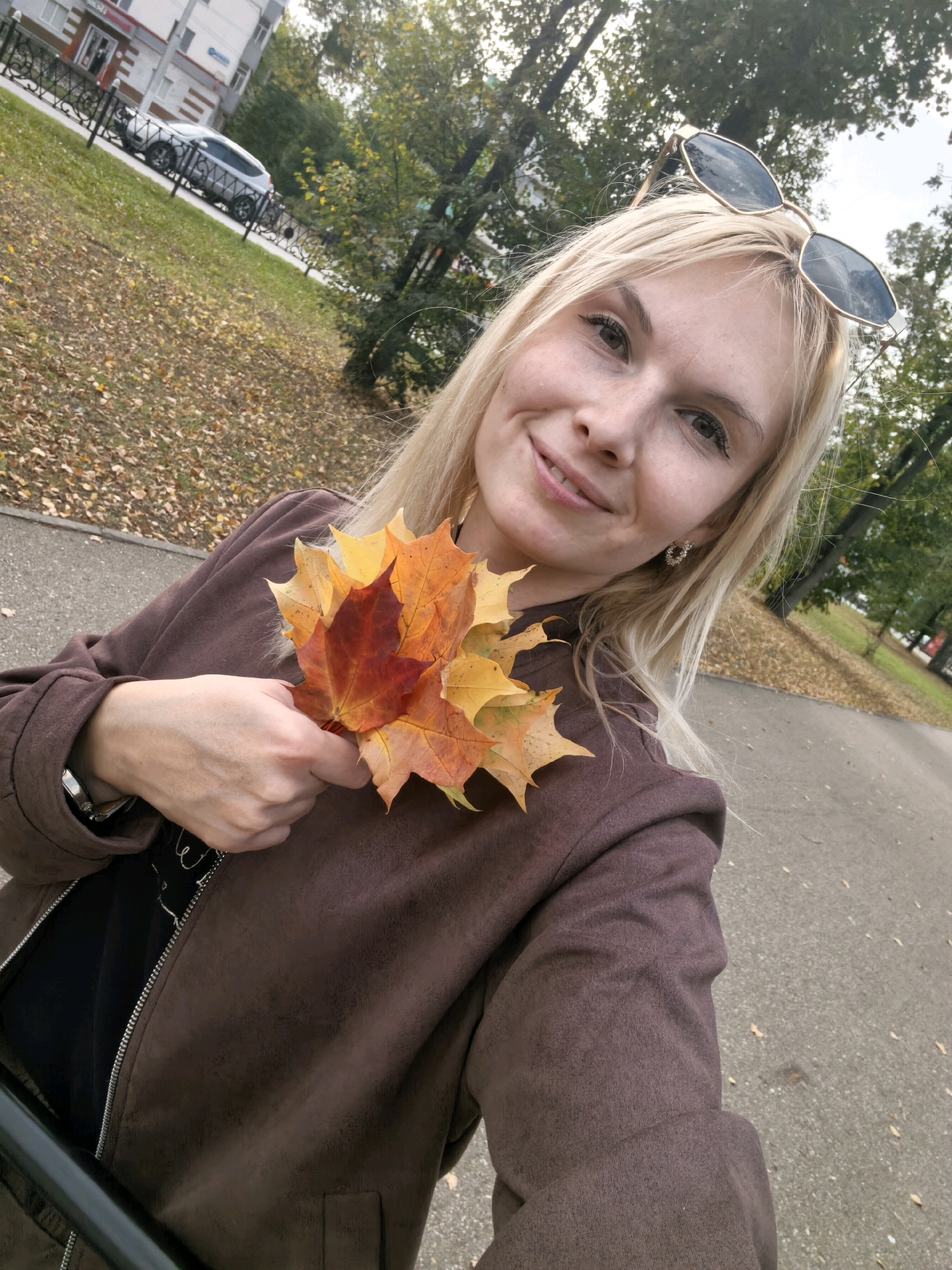 A blonde woman in a brown jacket and black shirt holds a bouquet of autumn leaves. She is outdoors in a park with trees and fallen leaves on the ground. A road is visible in the background, and a car is parked near a building on the left side of the image. The woman is smiling and looking at the camera. The sunglasses perched on her head suggest it is a sunny day. The location is in Sterlitamak, Russia, as indicated by the style of the building and the presence of a car common in that region.