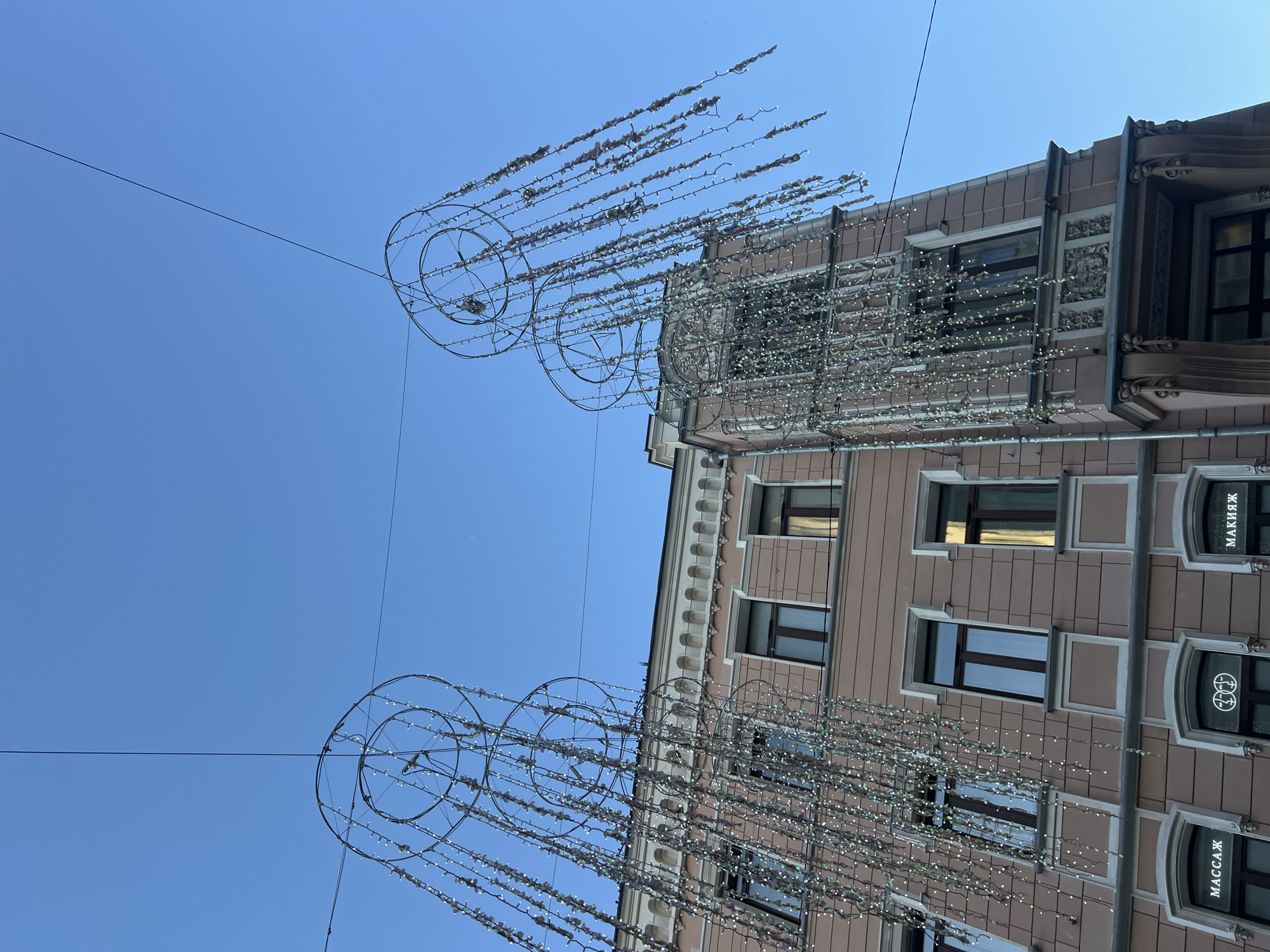 A low-angle exterior shot captures an urban scene featuring a building and elaborate street decorations against a clear blue sky in what is identified as Moscow, Russia.
The upper half of the image is dominated by a vibrant, cloudless blue sky, where a faint, thin crescent moon is visible just to the left of the image center. Several thin black wires crisscross the sky, suspending two large, circular metal decorative structures. These structures are adorned with numerous vertical strings of small, iridescent, bead-like lights or reflective elements, creating a cascading or waterfall effect. The lights appear to be inactive, consistent with daytime. The left decorative structure is more fully visible and closer to the foreground than the partially cropped right structure.
Below the suspended decorations, a multi-story building with a light rosy-beige or dusty pink facade and ornate white architectural trim occupies the lower half of the frame. The building features multiple rectangular windows with dark frames, some reflecting the blue sky. The top visible floor has a series of smaller arched decorative elements below the main cornice. The ground floor clearly displays three storefronts. From left to right, the windows feature white Cyrillic text on a dark background: "МАССАЖ" (MASSAGE), a circular logo depicting two stylized intertwined figures, and "МАКИЯЖ" (MAKEUP). The architecture suggests a historical urban setting, typical of central Moscow. No individuals are present in the scene.