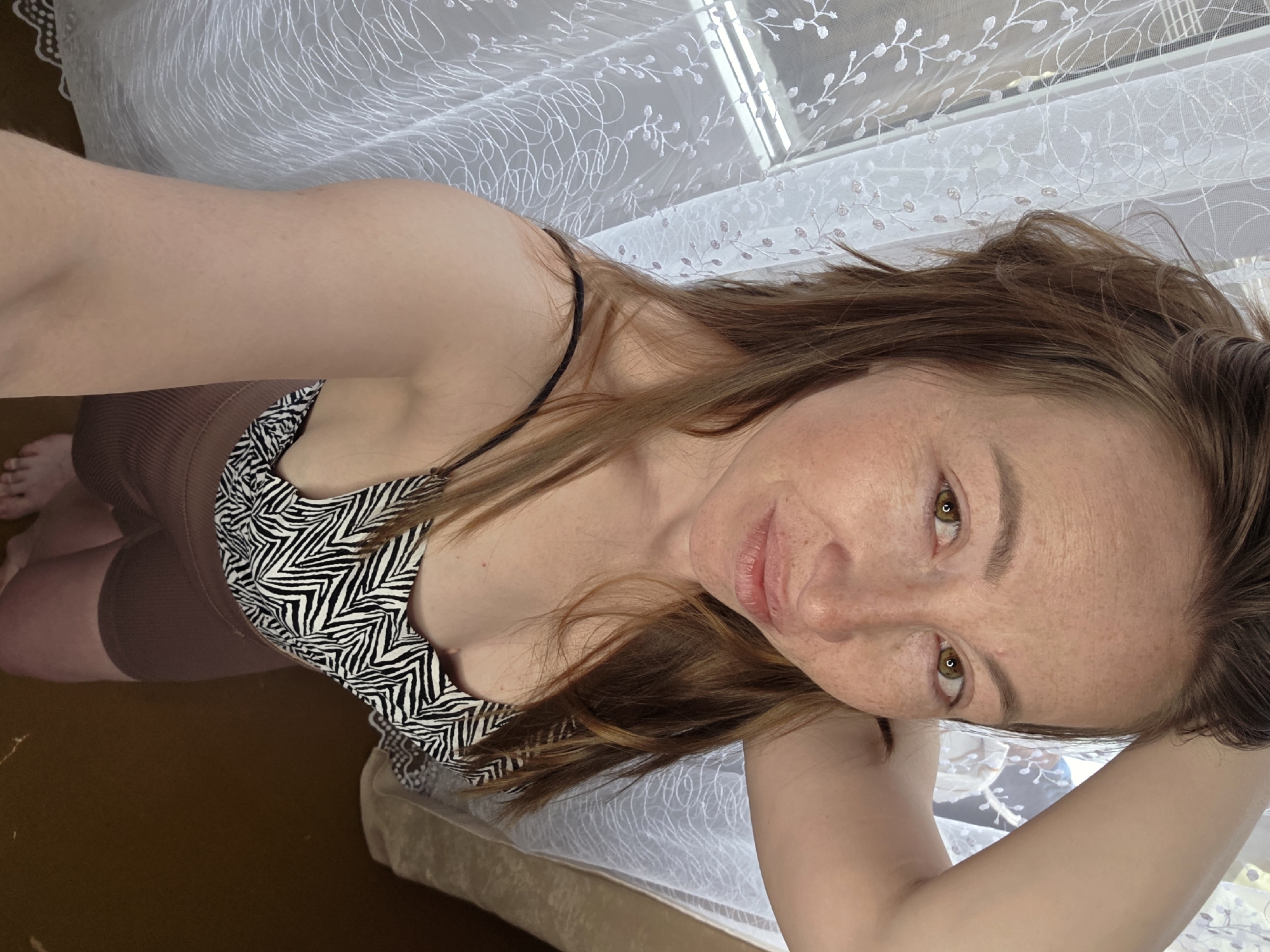 A woman is posing for a selfie indoors, in what might be her apartment, possibly in the city of Chaikovsky, Russia. She is wearing a zebra-print top with thin straps. Her hair is brown and shoulder-length, and she has freckles. The background shows a window covered with a sheer white patterned curtain. The floor is wooden, and there is part of a bed visible. She is holding her right arm behind her head.