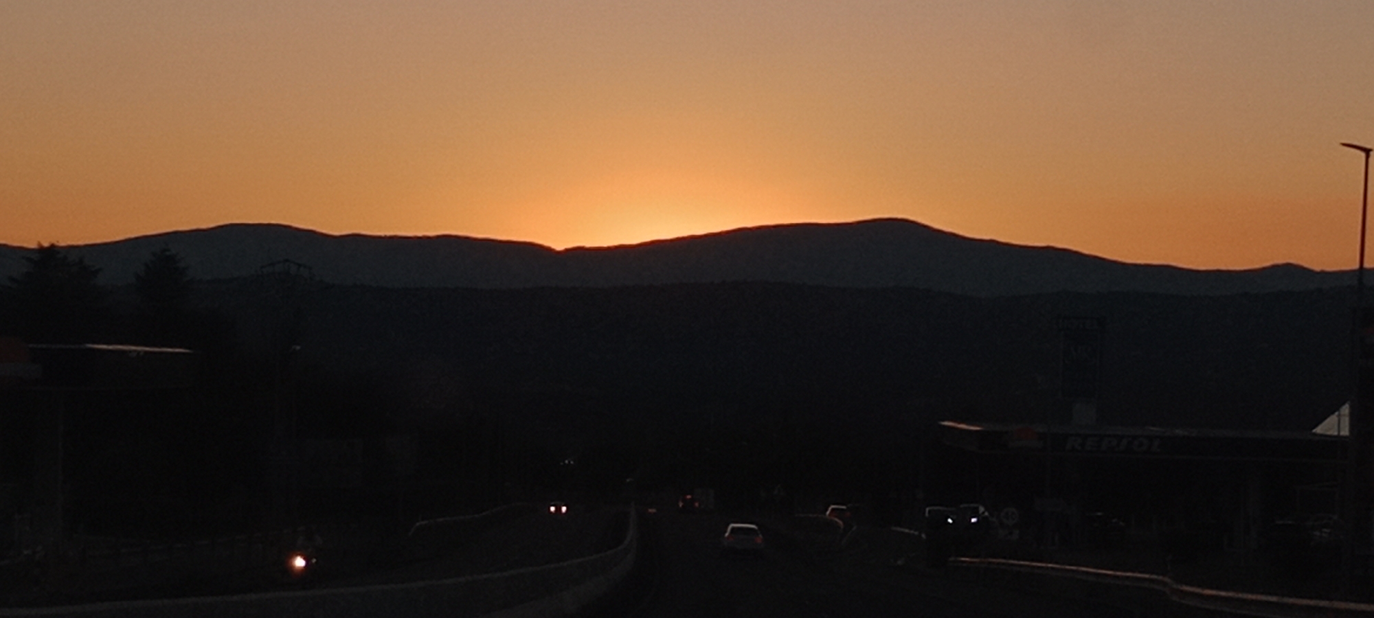 This image depicts a sunset over a mountainous landscape, likely near La Chopera, Spain. The sky is a vibrant gradient of orange and yellow, with the sun setting behind a dark silhouette of mountains in the distance. In the foreground, there is a dimly lit highway with several cars driving. On the right side of the image, a Repsol gas station is visible, also silhouetted against the fading light. The overall scene is one of twilight, with the natural beauty of the sunset juxtaposed with elements of human infrastructure.