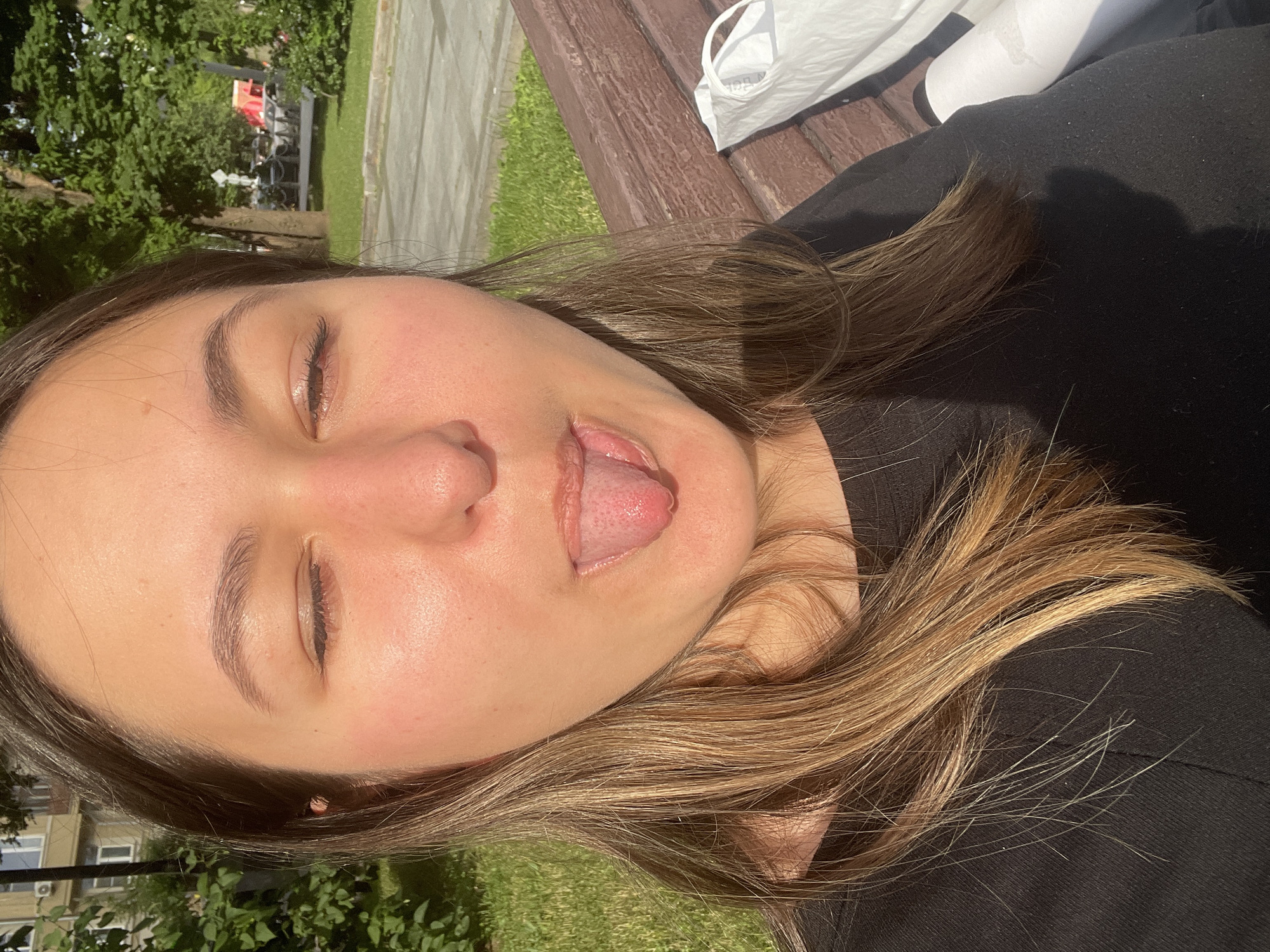 A young woman with light brown hair and closed eyes sticks her tongue out towards the camera, as she sits on a park bench. The background shows green grass, trees, and buildings, suggesting an outdoor urban setting.