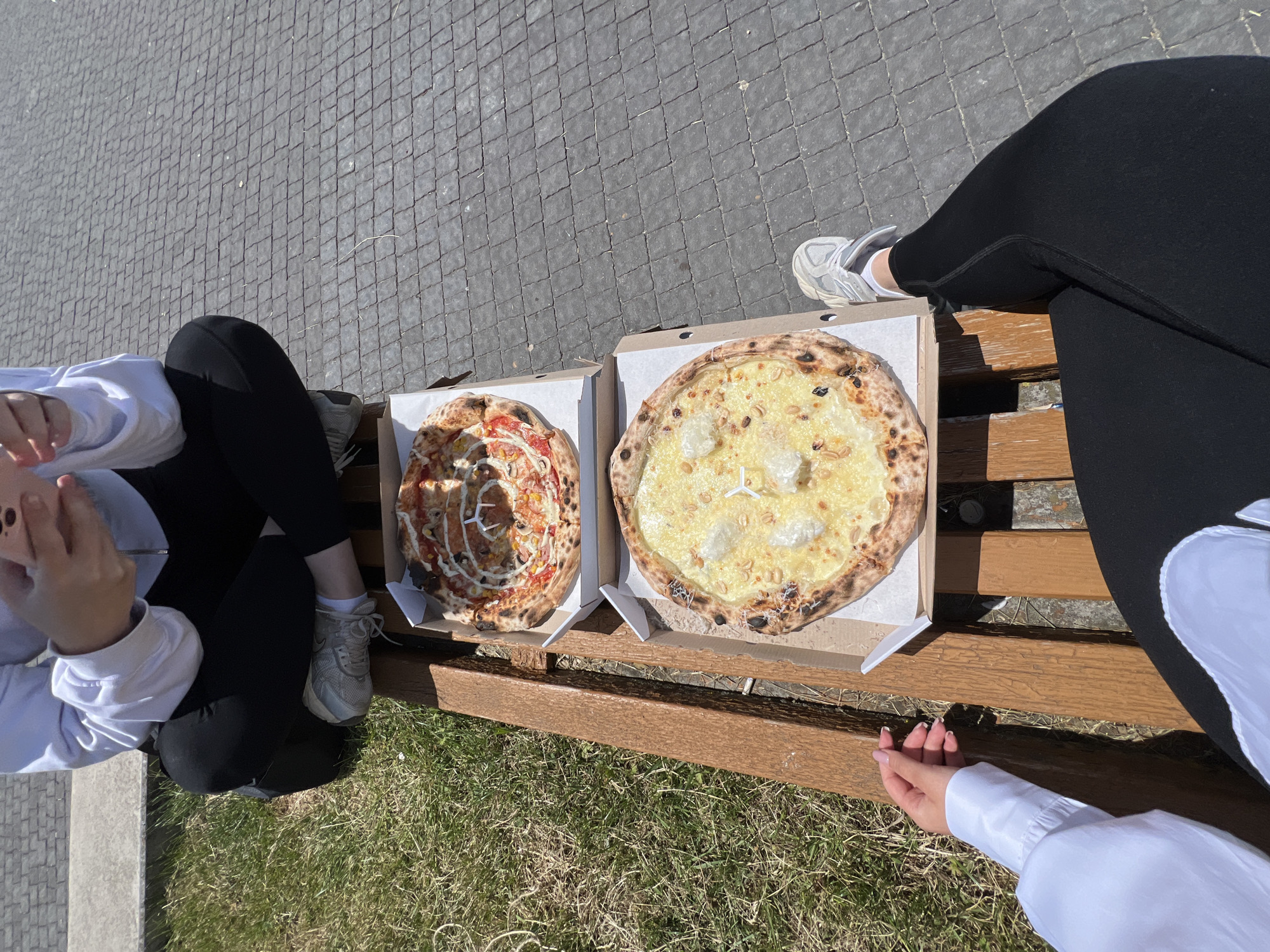Two individuals are seated on a wooden park bench on a sunny day. They are positioned at an angle, with their legs crossed or bent. The person on the left is wearing a white long-sleeved shirt and black pants. Their left hand, with manicured nails, rests on the bench near the pizza box. The person on the right is wearing black leggings and white sneakers. Both individuals are looking down at their phones.
In front of them, on the bench, are two open pizza boxes, each containing a pizza. The top pizza has a topping that appears to be a mixture of red sauce, white sauce, corn, and possibly mushrooms, arranged in a circular pattern. The bottom pizza is covered in melted cheese and appears to have some chunks of what could be ricotta cheese and possibly pine nuts.
The bench is situated outdoors, next to a patch of green grass. Behind the bench, the ground is paved with gray interlocking stones. The background shows a blurred view of the paved area.
The location is specified as Ternopil, Ukraine.