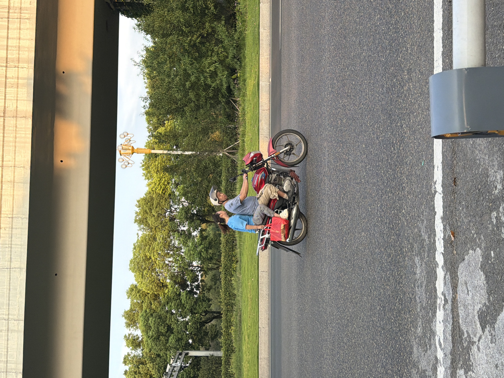 In Jiangjin, China, on a paved road with a white line marking, two individuals are riding a red motorcycle. The rider, wearing a helmet and a light-colored shirt, is looking ahead. The passenger, a woman with her hair tied back, wears a blue shirt and patterned pants, and she is looking at the rider. She has a red bag slung over her shoulder. The motorcycle has a silver rack on the back. The background features lush green trees and a yellow street lamp under a concrete overpass. The scene is lit by natural light, suggesting it is daytime.