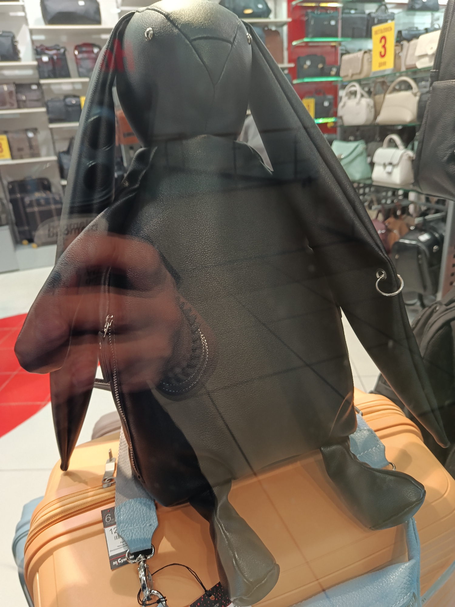 A black leather bunny-shaped backpack sits atop an orange suitcase inside what appears to be a store in Moscow, Russia. The backpack features long, dangling ears, one of which has a silver ring. The suitcase has a blue strap attached, and a price tag with Cyrillic text is visible. The store has shelves stocked with various handbags in the background, indicating it might be a bag or luggage store. A reflection of a person's hand and forearm is visible on the glass of the store window.
