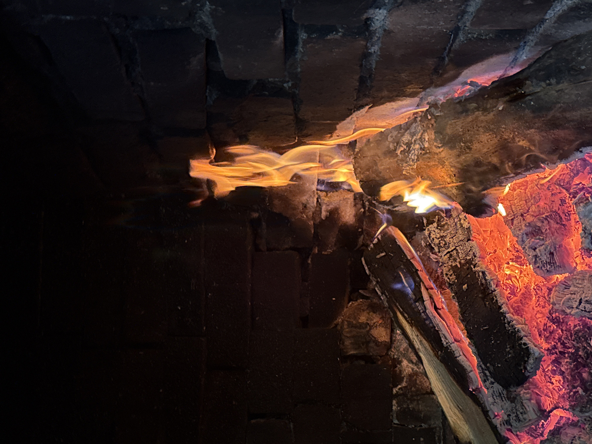 A close-up shot of a fire burning inside a brick fireplace. Flames are visible between the logs, and embers glow within the wood. The bricks of the fireplace appear dark. The location in the city of Lilkovo, Bulgaria, is not discernible from the image.