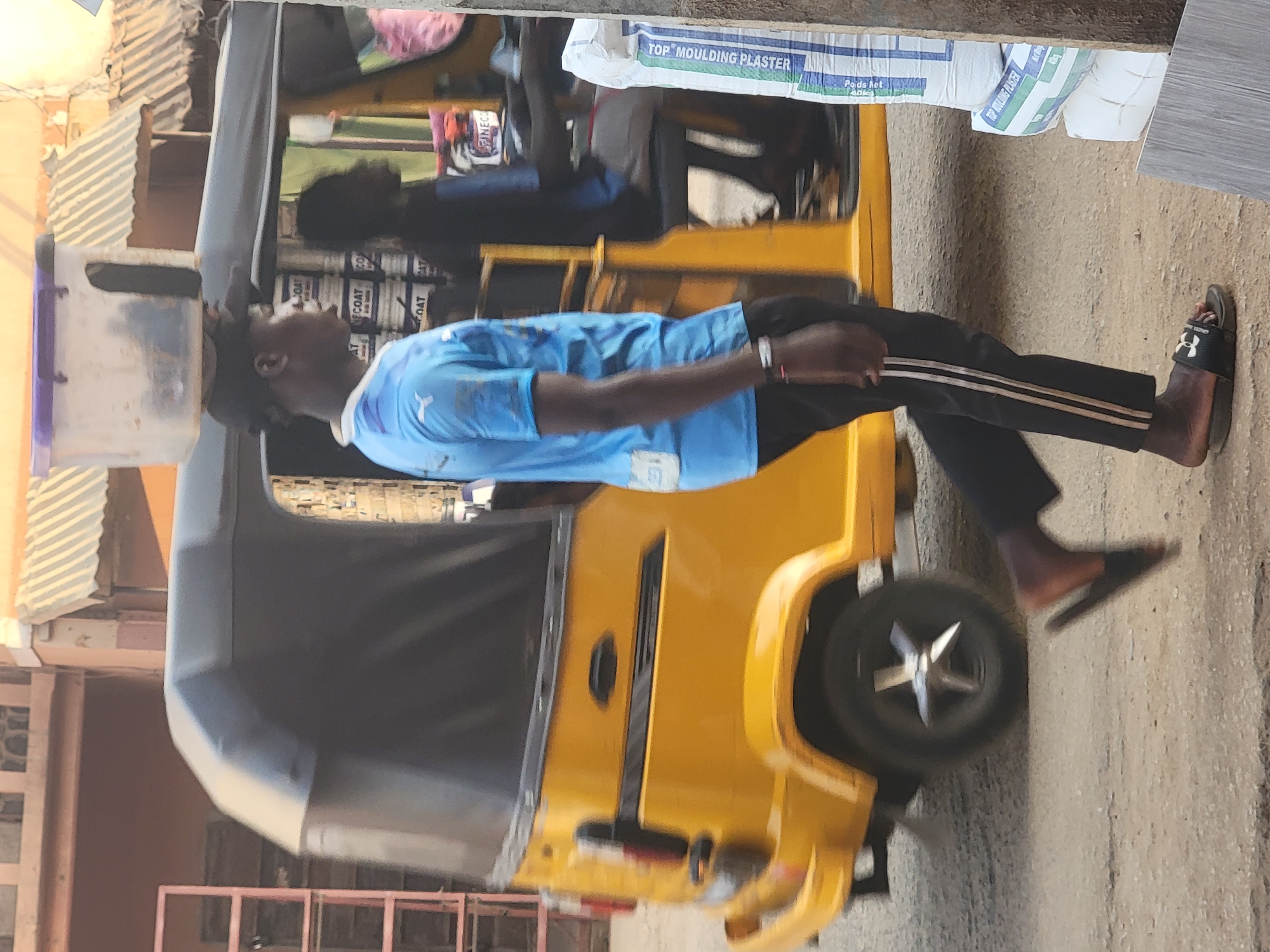 The image depicts a man standing next to a yellow tricycle (auto-rickshaw) on a street in Maiburiji, Nigeria. The man wears a light blue t-shirt, black pants with white stripes, and sandals. He appears to be looking towards the tricycle, which has a container on top. A person is seated inside the tricycle. Behind the tricycle are bags of moulding plaster. The street appears to be paved, and there are buildings and structures in the background.