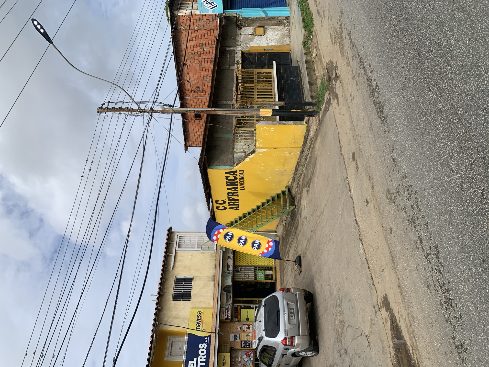 The image captures a street scene in La Tagua, Venezuela, featuring a row of businesses along a roadside. In the foreground, a silver SUV is parked on the paved street, which shows signs of wear and tear with cracks and patches. Adjacent to the street is a dirt and asphalt area.
To the left, a yellow building houses several businesses. One banner reads "EL TROS C.A." and another displays "mavesa." A blue flag with "PAN" repeated in yellow circles is prominently displayed on a stand in front of this building, alongside a green staircase leading to an upper level.
The middle section of the image is dominated by a brightly painted yellow wall with the text "CC ARFRANCA LA VECINDAD." A metal staircase with green railings ascends this wall. This section appears to be a more dilapidated commercial structure with exposed concrete and brickwork, featuring barred windows and a dark-colored doorway.
To the right, a building with a red brick facade is visible, with a section of it appearing to be unfinished or in disrepair. Further to the right, a storefront with a bright blue shutter is partially visible. The sky is overcast with clouds. Overhead, a complex network of power lines crisscrosses the scene, supported by a wooden pole. A modern streetlamp with three lights extends from another pole on the right side of the image.