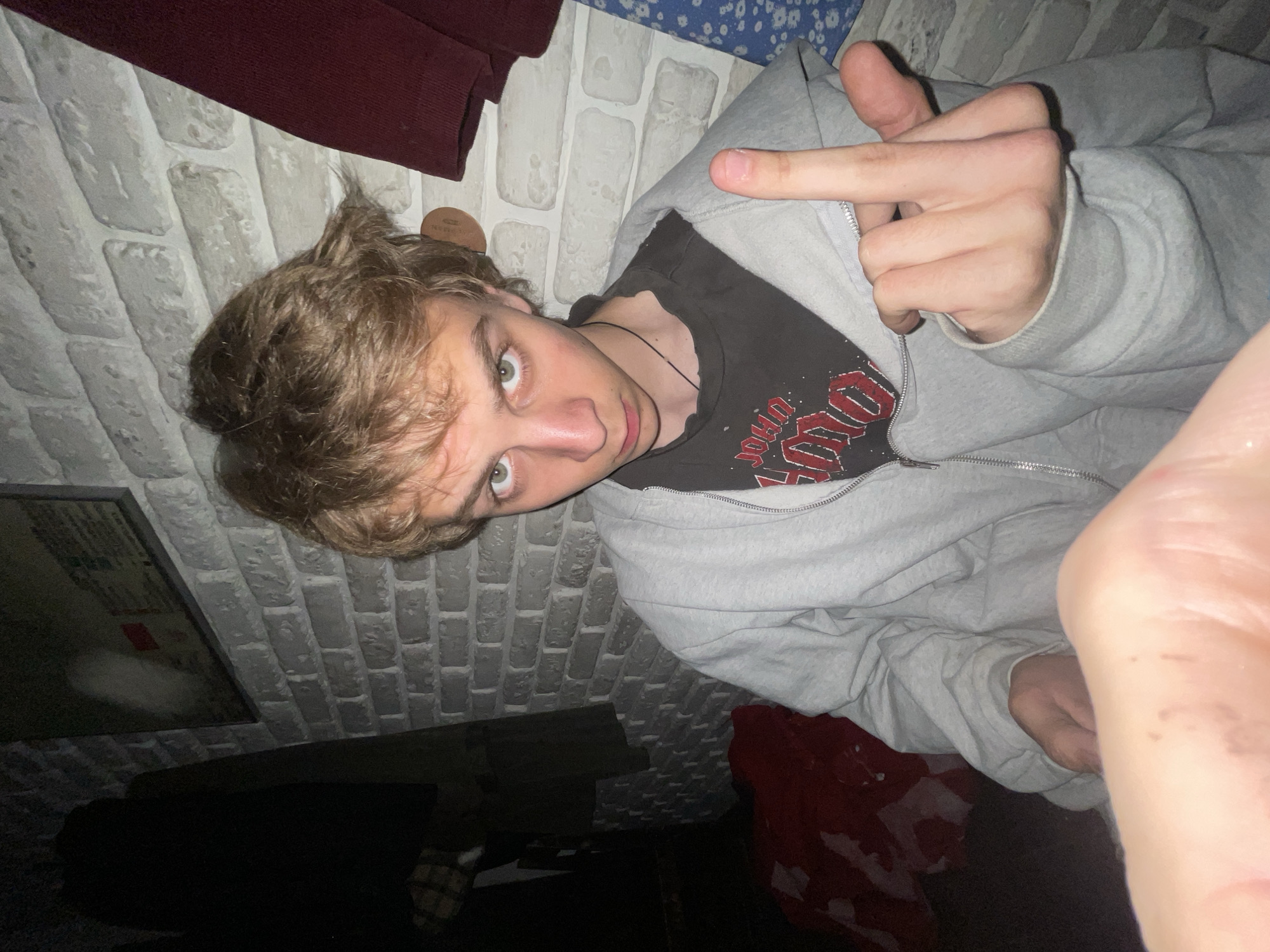 A young man with blond hair, wearing a grey hooded sweatshirt and a black t-shirt, is lying down and extending his middle finger towards the camera. The background includes a brick-patterned wall, a maroon-colored garment, and a dark, indistinct area. The location is not discernible from the image. Moscow, Russia, cannot be confirmed.