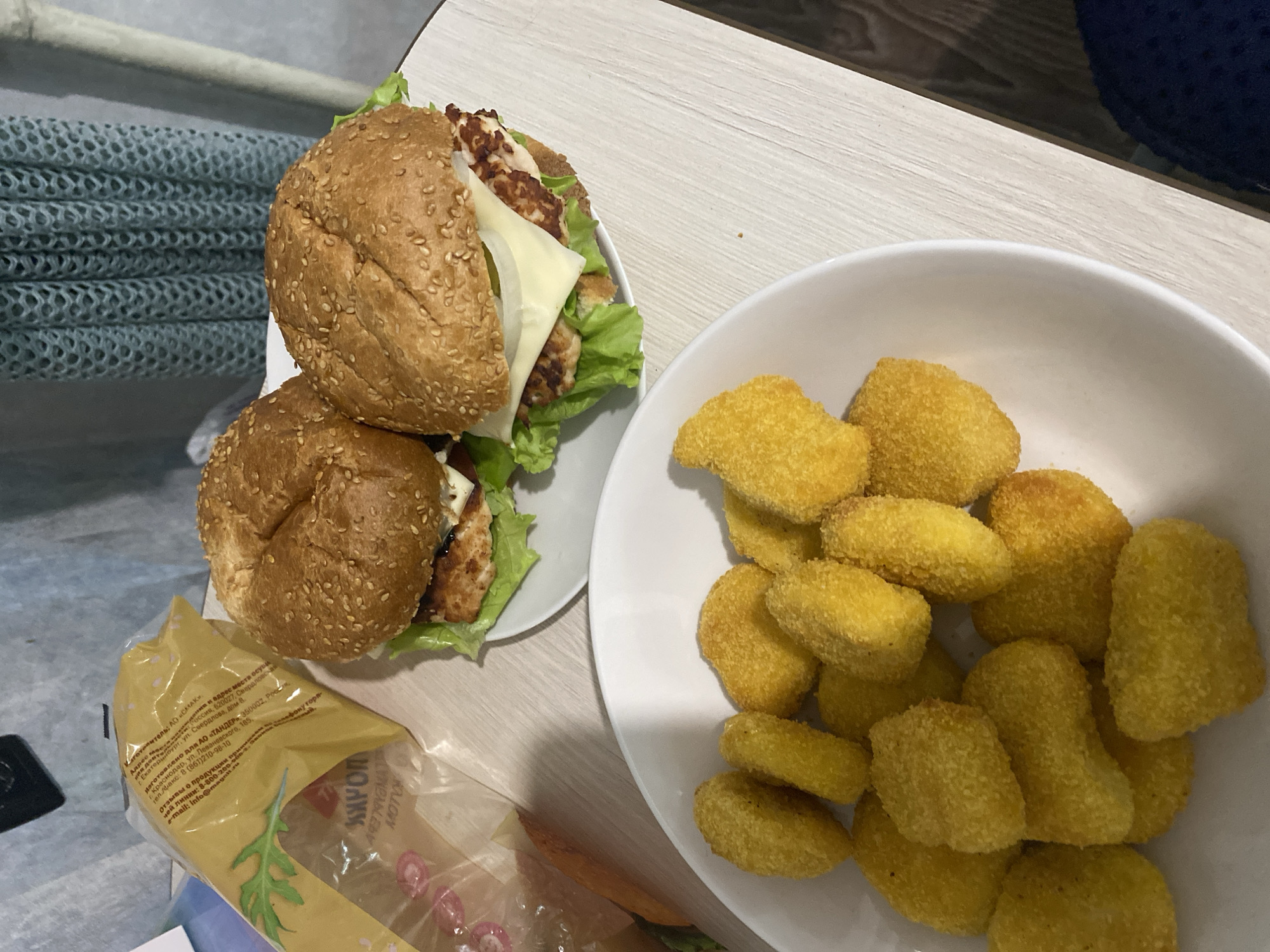 The image shows a tabletop setting with food items. Two sesame seed burgers, stacked on top of each other, are positioned on a small white plate. Lettuce and a slice of cheese are visible within the burgers. Next to the plate is a white bowl filled with numerous golden-brown chicken nuggets. Adjacent to the burgers, there is a partially visible opened package. The text on the package is in Russian, indicating that the manufacturer is AO "SMAK" with location in Russia, 620027, Sverdlovsk region, Ekaterinburg, Sverdlova street, 8. A portion of a gray textured surface is visible to the left of the food items. The tabletop appears to be a light wood-grain surface. There is no evidence to place this image in Izhevsk, Russia.