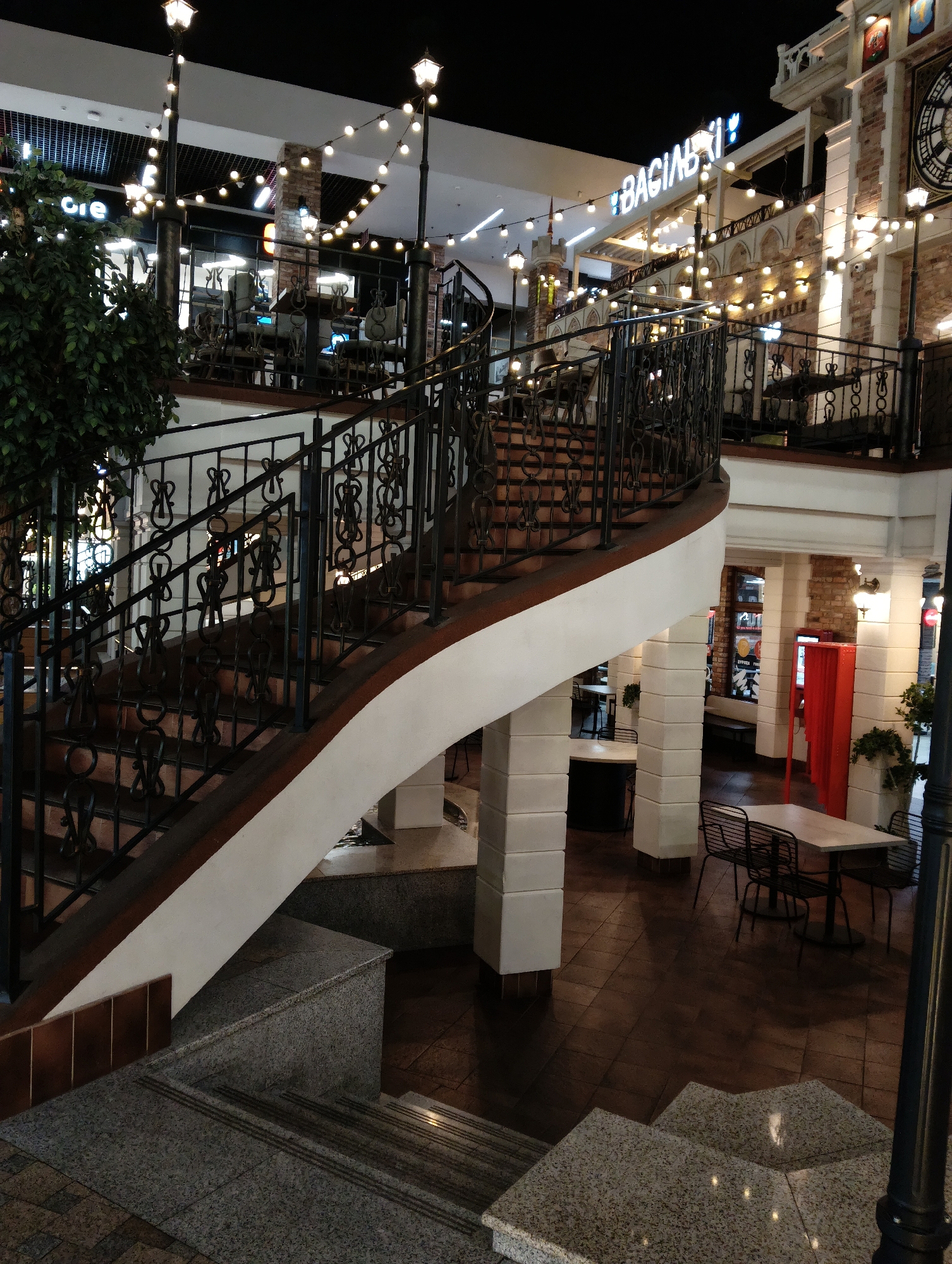The image shows an indoor scene, likely a mall or a restaurant, featuring a prominent spiral staircase with ornate black metal railings. The staircase is illuminated by string lights and street lamps. To the right of the staircase, there are tables and chairs set up, suggesting a dining area. A large red kiosk-like structure is also visible. In the background, there is a sign that reads "BAGINSKI," with some Cyrillic lettering above it. The overall ambiance is that of a well-lit, modern establishment with a touch of vintage or industrial design elements. This establishment is identified as Baginski in Hrodna, Belarus. There are no people visible in the image.