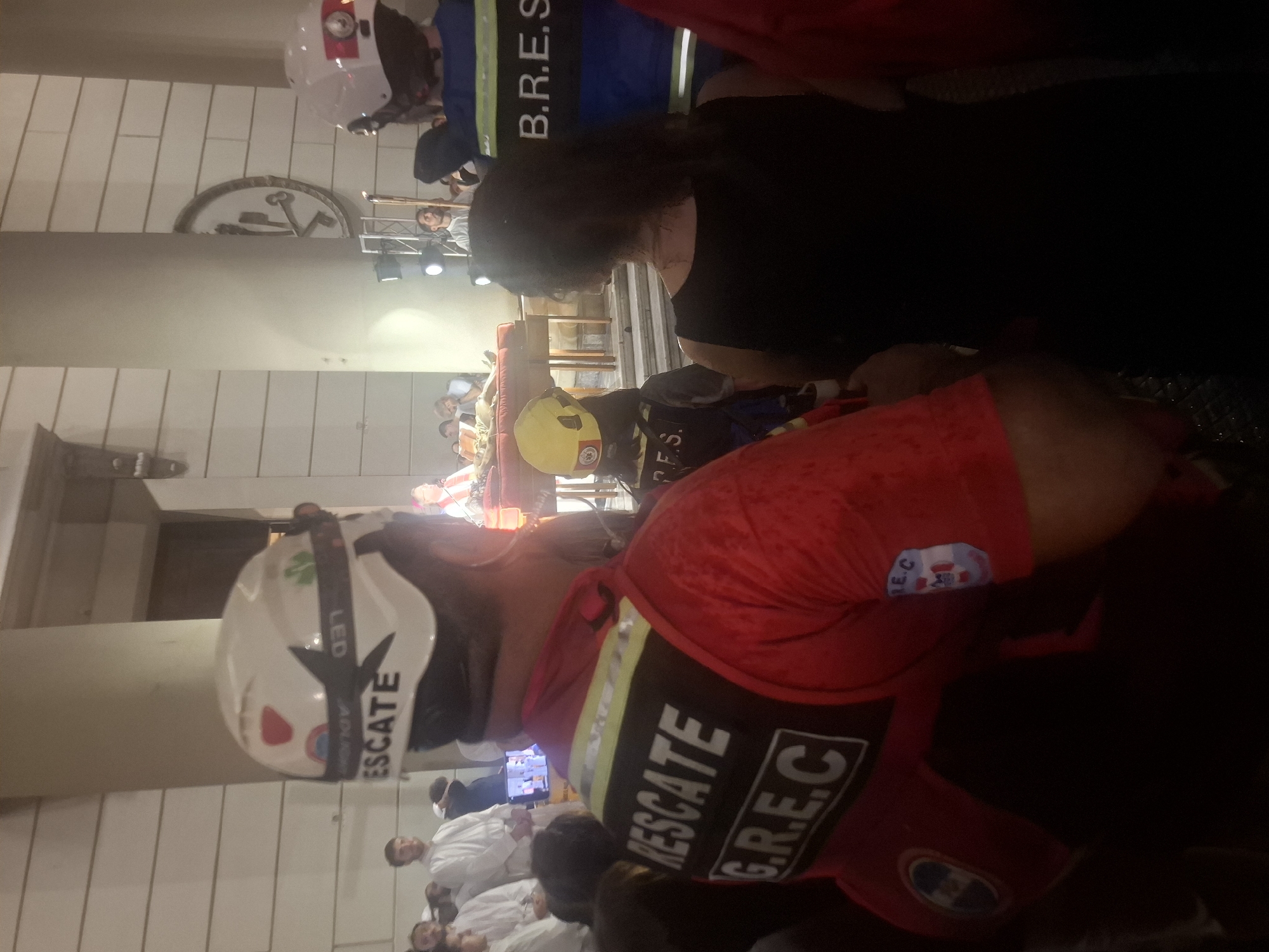 The image depicts a scene in Buenos Aires, Argentina, featuring a rescue team ("RESCATE G.R.E.C") in red uniforms and helmets, with one helmet clearly showing the word "RESCATE." People in white uniforms are present in the background, possibly as part of a ceremony or event. There are columns and architectural details suggesting a public building in the city center. The illumination suggests a nighttime setting or an indoor location with artificial lighting. The presence of emergency personnel indicates a staged rescue scenario or a demonstration.