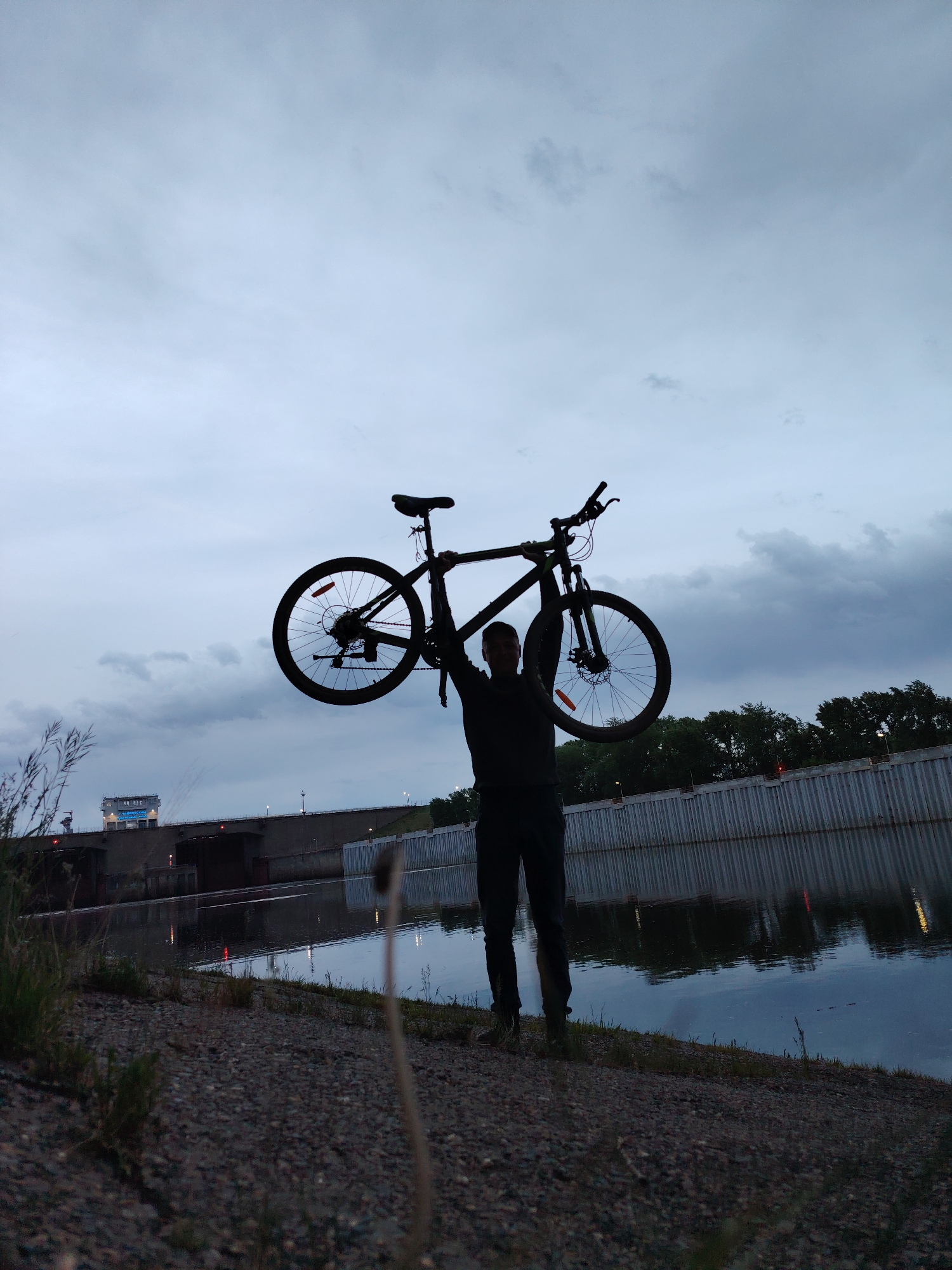 A person silhouetted against a twilight sky holds a bicycle aloft on the bank of a body of water. The person appears to be standing on a gravelly bank next to the water. In the background, a concrete structure with a building on top spans the water. Additional structures line the bank on the other side. The scene appears to be in Chaikovsky, Russia, based on the built structures in the background.
