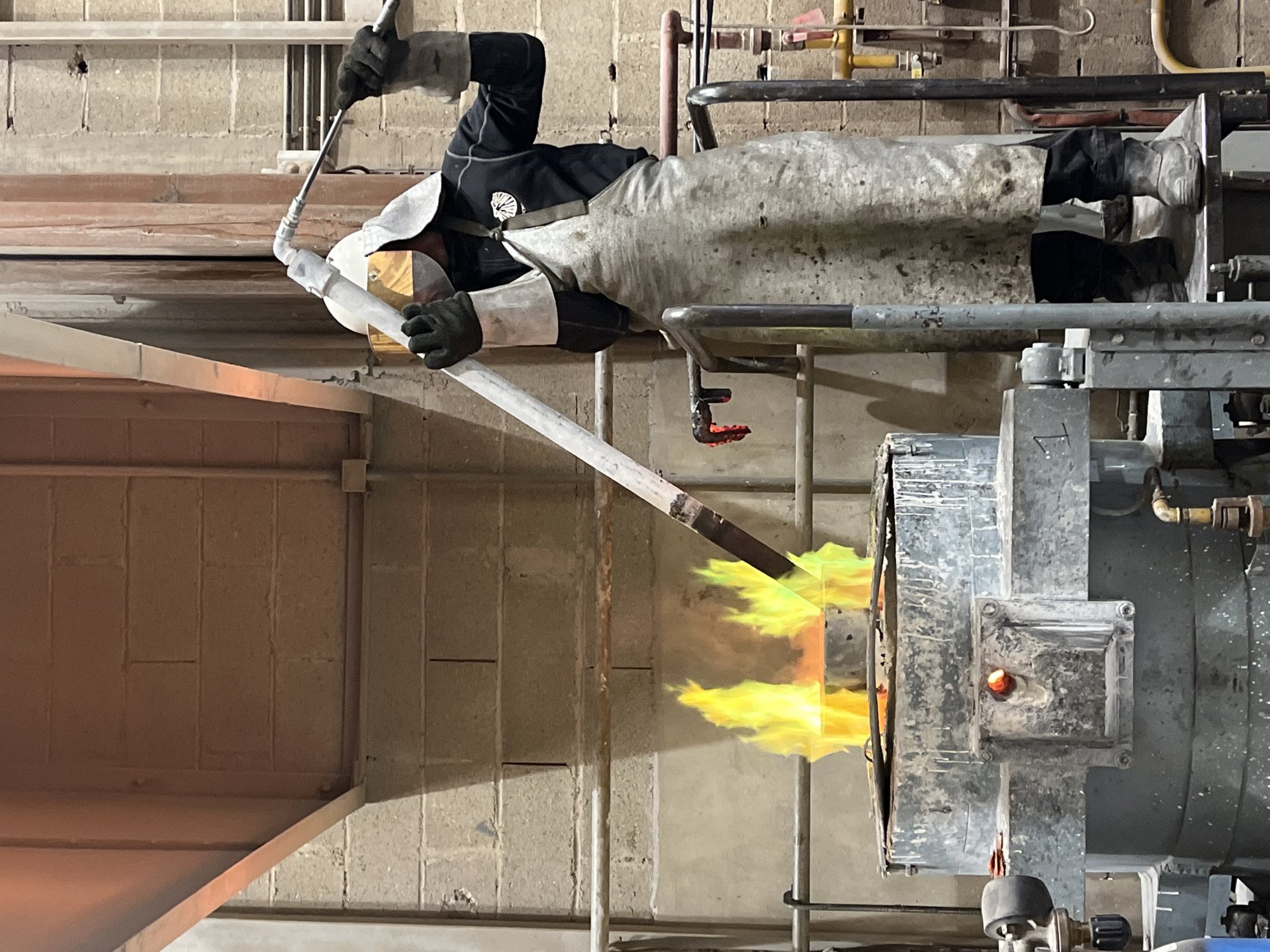 In an industrial or workshop setting, a person clad in protective gear is actively engaged in a high-temperature operation. The individual wears a dark long-sleeved shirt, a heavy protective apron, gloves, work pants, boots, and a face shield/visor, indicating work with hazardous materials or extreme heat.
The central activity shows the person holding a long metal rod or blowpipe, which extends into the flaming opening of a large industrial furnace or kiln. Bright yellow and orange flames are vigorously emanating from this opening, highlighting the intense heat involved in the process, which could be glassblowing, metal smelting, or a similar high-temperature craft.
The background features concrete block walls and a network of various metal pipes and structural elements, suggesting a functional, working environment. The lighting is internal, as expected in such a facility. The person's posture conveys focus and dedication to the task. No specific time of day or external weather conditions are discernible, and no legible text is visible in the frame. The location context provided is Saint-Rémy-lès-Chevreuse, France, though this is not visually confirmed by the image itself.