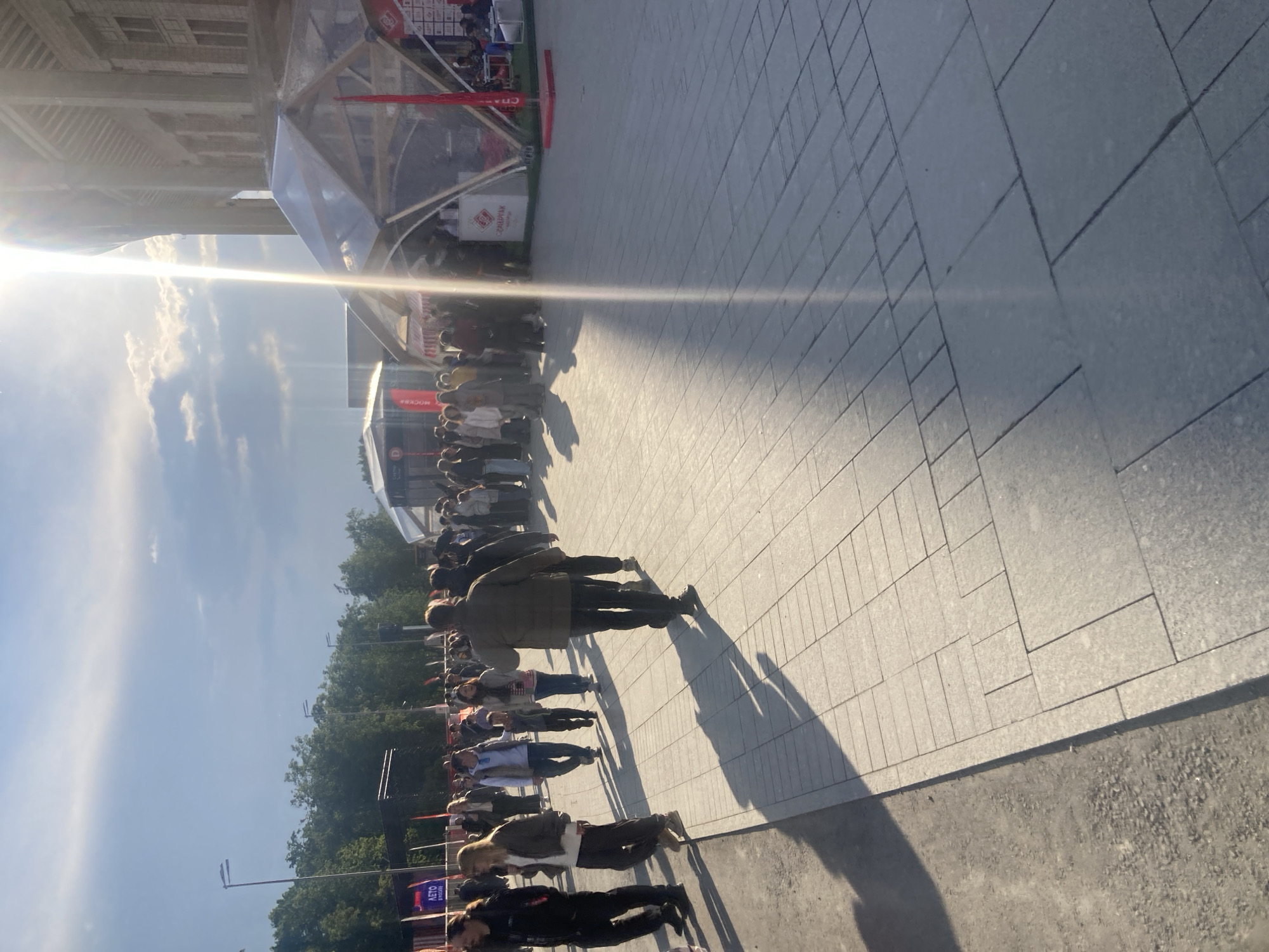 The image shows a line of people waiting outside a structure. The scene is well-lit with strong sunlight. The people are standing on a paved area that appears to be made of large stone tiles. The structure is partially visible, with a clear, dome-like section on top. A building is also visible in the background.
A sign is visible reading "MOCKBA." The location is Moscow, Russia.