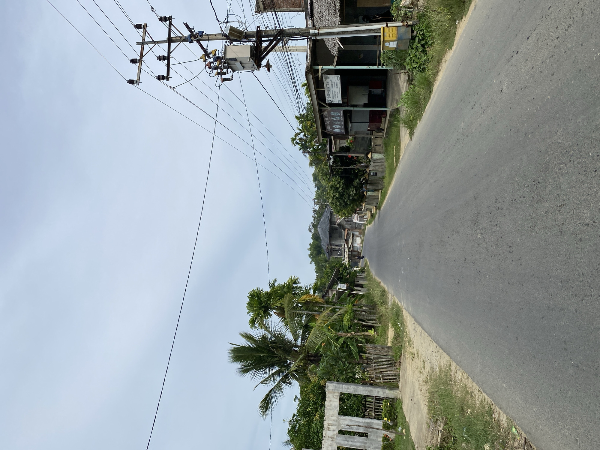 This media file depicts a roadside scene in Lhokseumawe, Indonesia. The image is oriented vertically, with the road running diagonally from the bottom right to the top left. The sky is overcast, suggesting a cloudy day.
On the left side of the road, there is a utility pole with numerous wires and a transformer, indicating an active electrical infrastructure. Adjacent to the pole are several small buildings with corrugated metal roofs, appearing to be shops or small businesses. One sign visible on a building reads "DARUHINTAN" followed by smaller text that is difficult to decipher. Further down the road, more buildings and vegetation are visible.
The right side of the image is dominated by the asphalt road, with a grassy and sandy verge along its edge. Further to the right of the verge, there are more trees, including palm trees, and some simple fences. The overall impression is of a rural or semi-urban setting in a tropical or subtropical climate. No people are clearly visible in the frame. The time of day appears to be daytime, given the general illumination. The scene is static, with no specific activity or event taking place.