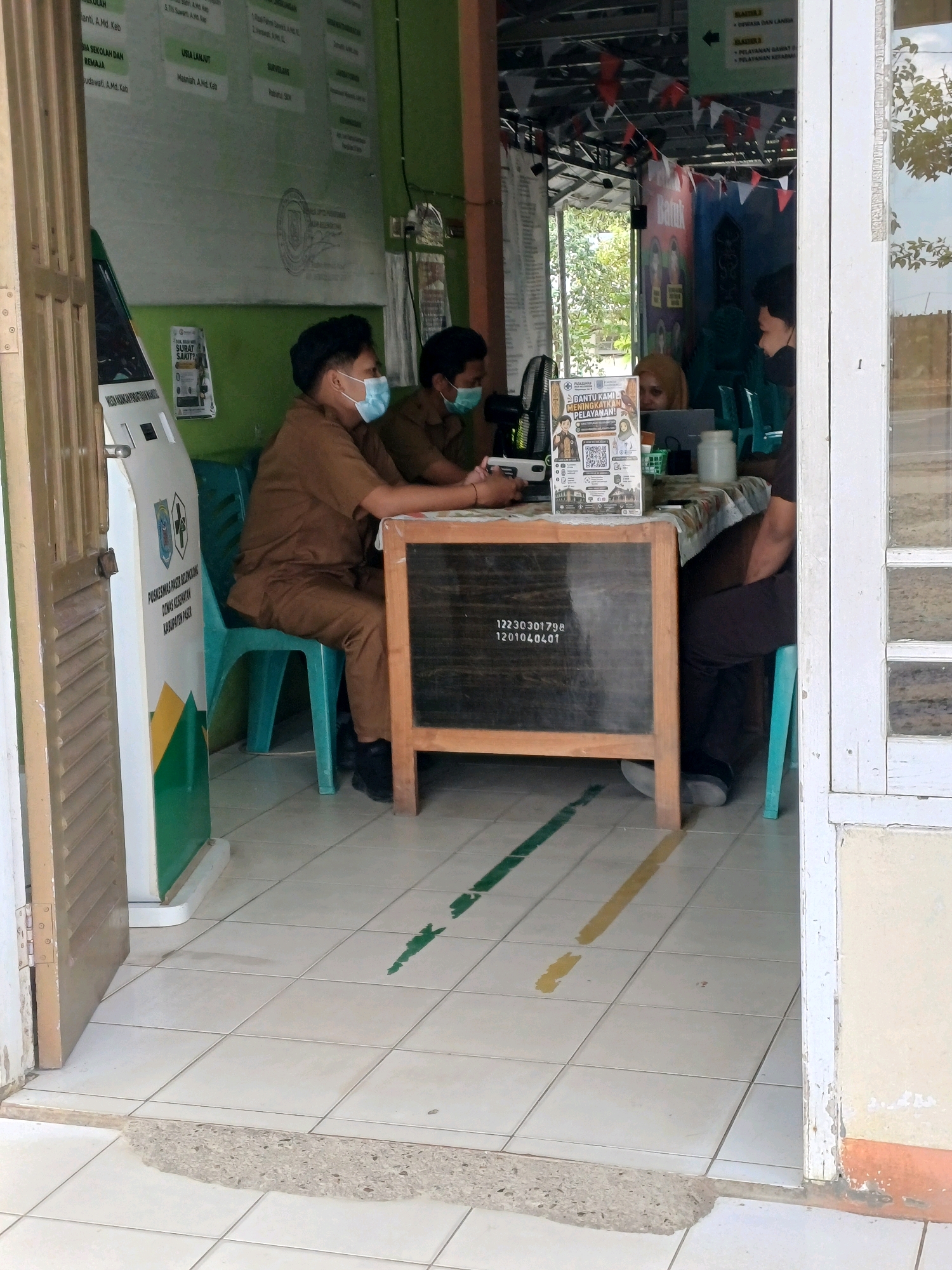 In Pasir Belengkong, Indonesia, an indoor scene shows three individuals wearing face masks and brown uniforms seated behind a table. Two individuals are facing forward, one appears to be looking at a laptop. The third individual is seated at the table, facing away from the camera. On the table, a laptop, a white jar, and a green basket are visible. A chalkboard with white text and numbers is placed in front of the table. To the left of the table, a white kiosk with green and yellow accents is positioned near a closed wooden door. The floor is tiled, with green and yellow lines painted on it, possibly indicating social distancing markers. The background features a green wall with posters and a metal ceiling with decorative red and white flags.