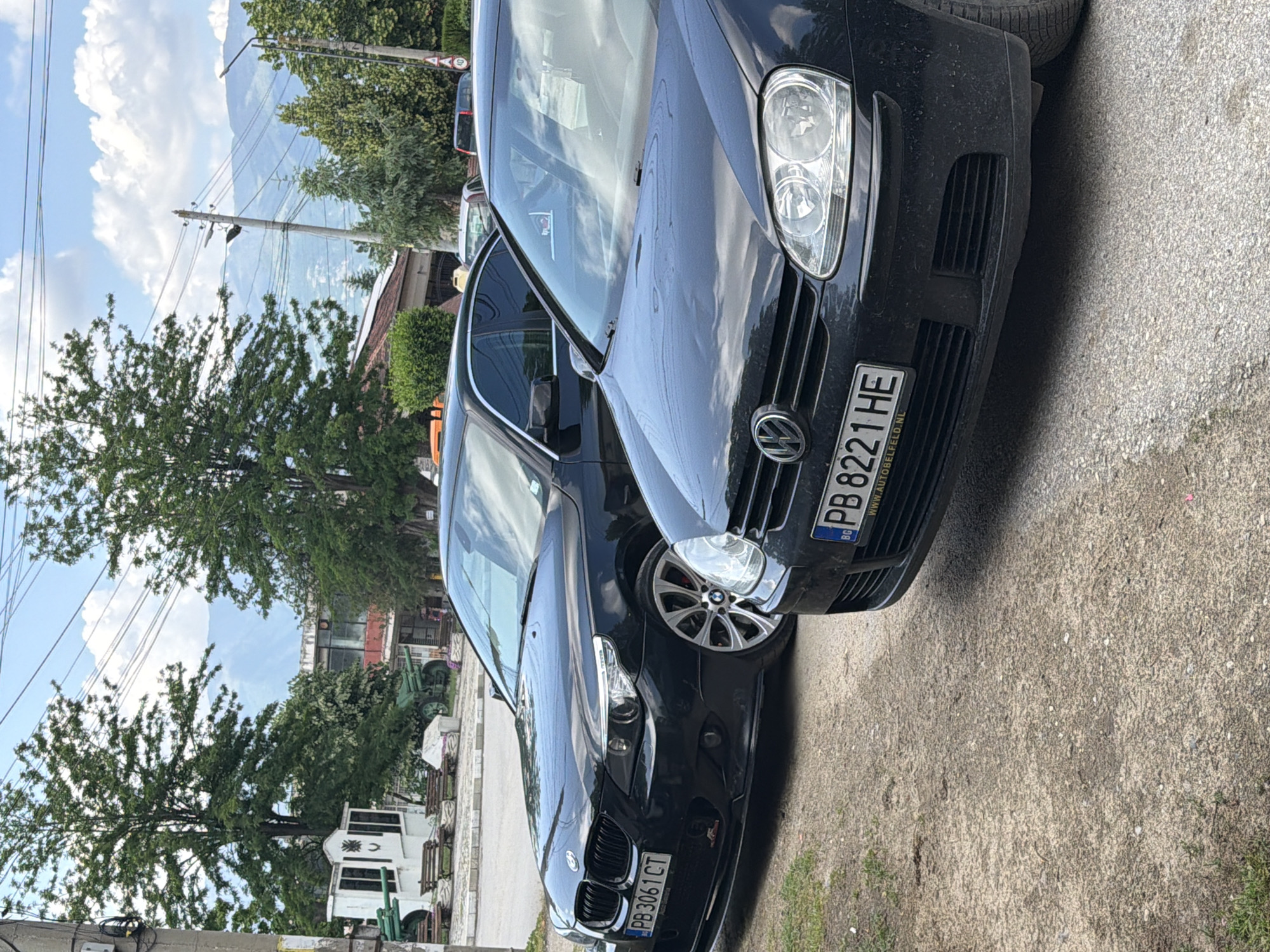 This image captures an outdoor scene in Dabene, Bulgaria, under a partially cloudy sky during daylight hours. In the immediate foreground, two black passenger vehicles are parked on an unpaved, gravelly surface. The car on the left is a black BMW 5-Series (E60/E61 generation) with a Bulgarian license plate "PB 3061 CT," featuring multi-spoke alloy wheels and a modified front splitter. The car on the right is a black Volkswagen Golf (Mk5 generation) with a Bulgarian license plate "PB 8221 HE," which includes a frame displaying "WWW.AUTOBELFELD.NL."
Behind the vehicles, on the left, a white memorial structure is visible, flanked by two green artillery cannons positioned on a paved public square. Several wooden benches are arranged around this memorial area. Large deciduous trees frame this section of the scene. To the right of the memorial, various buildings are visible, including one with solar panels on its roof. Utility poles with multiple overhead power lines traverse the mid-ground. A circular speed limit sign indicating "40" km/h is partially visible on the far right. In the distant background, hazy mountains rise beneath the clouds. No people are visible in the scene.