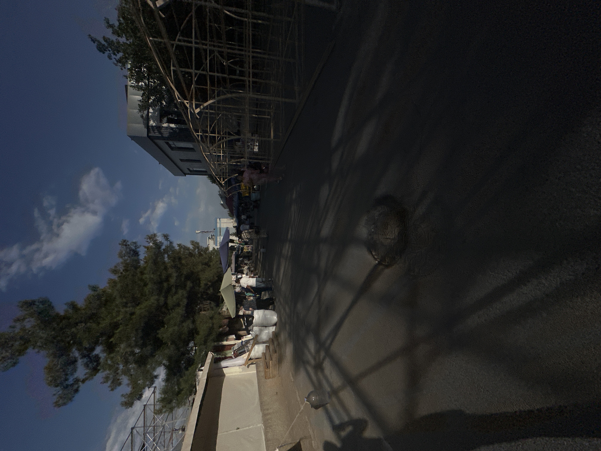 The image depicts an outdoor scene in Tashkent, Uzbekistan, likely a market or street scene. The perspective is tilted, with the sky and some buildings on the left and the ground and shadows on the right.
In the foreground on the left, a tall green tree with dense foliage is prominent, with a building in the background partially obscured by the tree and a metal scaffolding structure. Further down the street, there are several people visible, some appearing to be vendors or shoppers at stalls. Umbrellas, bags, and what appear to be goods are present. The ground is asphalt, with distinct shadows cast by the scaffolding and possibly trees. A circular manhole cover is visible on the pavement.
The time of day appears to be late afternoon or early evening, indicated by the long shadows and the lighting conditions. The sky is a deep blue with some scattered white clouds. The overall mood seems to be bustling, with activity suggested by the presence of multiple people and what look like commercial setups. There is no visible text in the image.