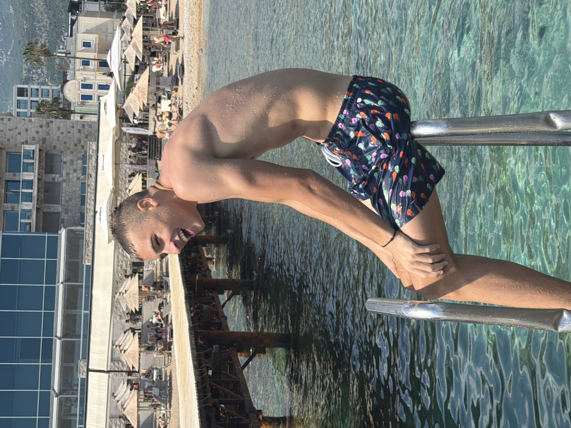 A young man, wet and with sweat glistening on his skin, is captured in mid-action, bending over a metal railing. He wears dark swim trunks with a colorful, patterned design. His face is contorted in a grimace of effort or excitement as he looks towards the viewer, his eyes closed and mouth open. His hands grip the railing, and his body is arched, suggesting he is either jumping into or emerging from the water.
The scene is set at a beach resort in Budva, Montenegro. In the background, a beach is visible with rows of sun loungers and umbrellas, indicating a sunny day. Buildings constructed with stone and glass are also present, hinting at the modern infrastructure of the resort. The water is clear and turquoise, reflecting the bright sunlight. The overall impression is one of a vibrant vacation setting, with the young man's energetic pose embodying the spirit of summer recreation. There is no visible text in the image.