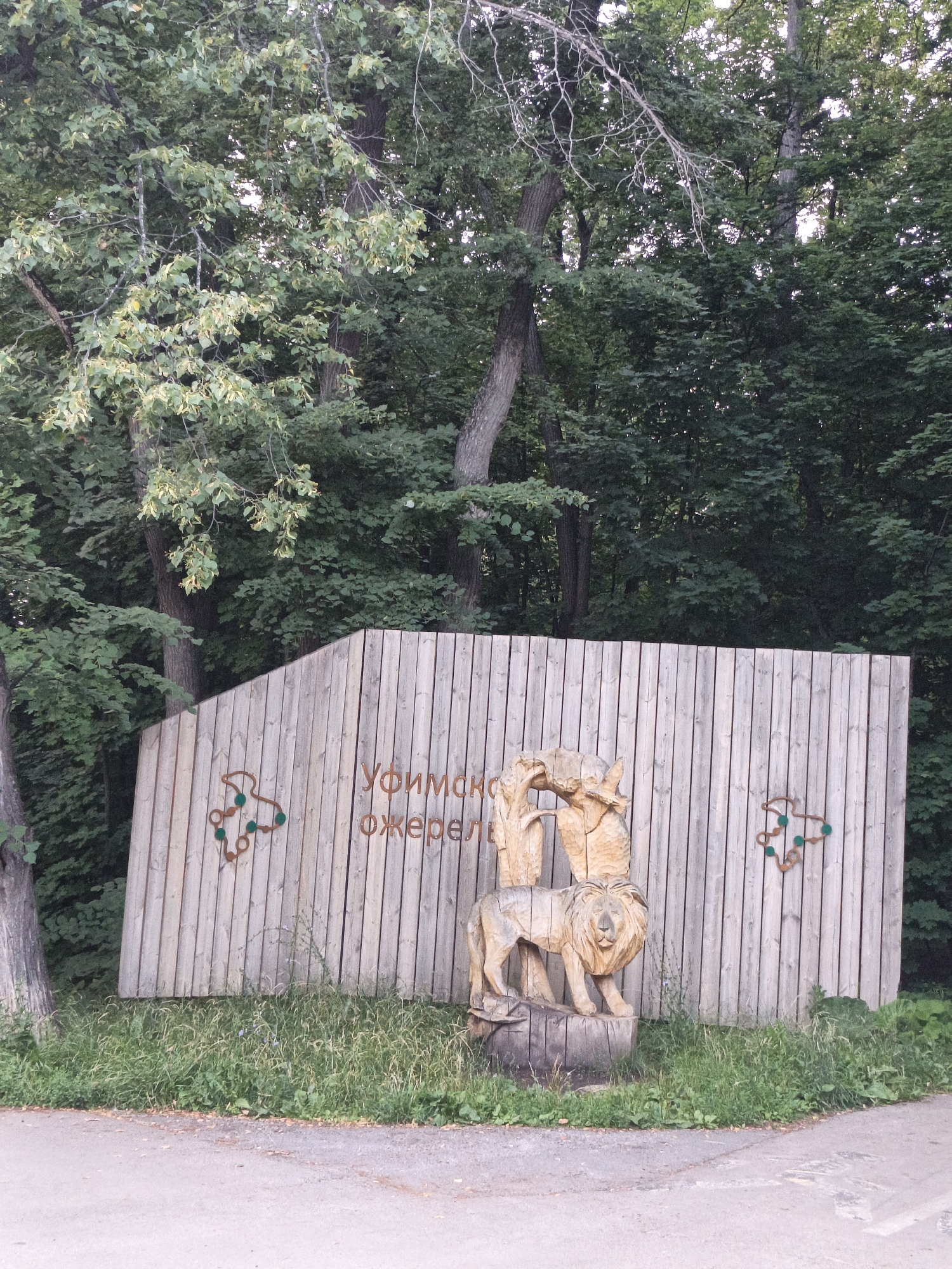 This is a medium shot, eye-level, outdoor, daytime photograph of a wooden fence with carvings at the Ufimsky Dzherel (Ufa Source) natural monument. The fence is made of vertical wooden planks and curves to the right. To the left of center on the fence, there is text carved in Cyrillic that reads "Уфимск(ий)" above "джерел". To the left and right of the text are decorative metallic cutouts shaped like abstract trees with green circular embellishments. In the center of the fence, there is a wooden carving of two figures embracing above a carving of a lion standing on a tree stump. The lion carving is detailed, showing its mane and facial features. The fence is set against a backdrop of dense green foliage and trees, suggesting a natural or park-like setting. In the foreground, there is a dirt path and grass with some small plants. The lighting suggests it is overcast or late afternoon. There are no people visible in the image.