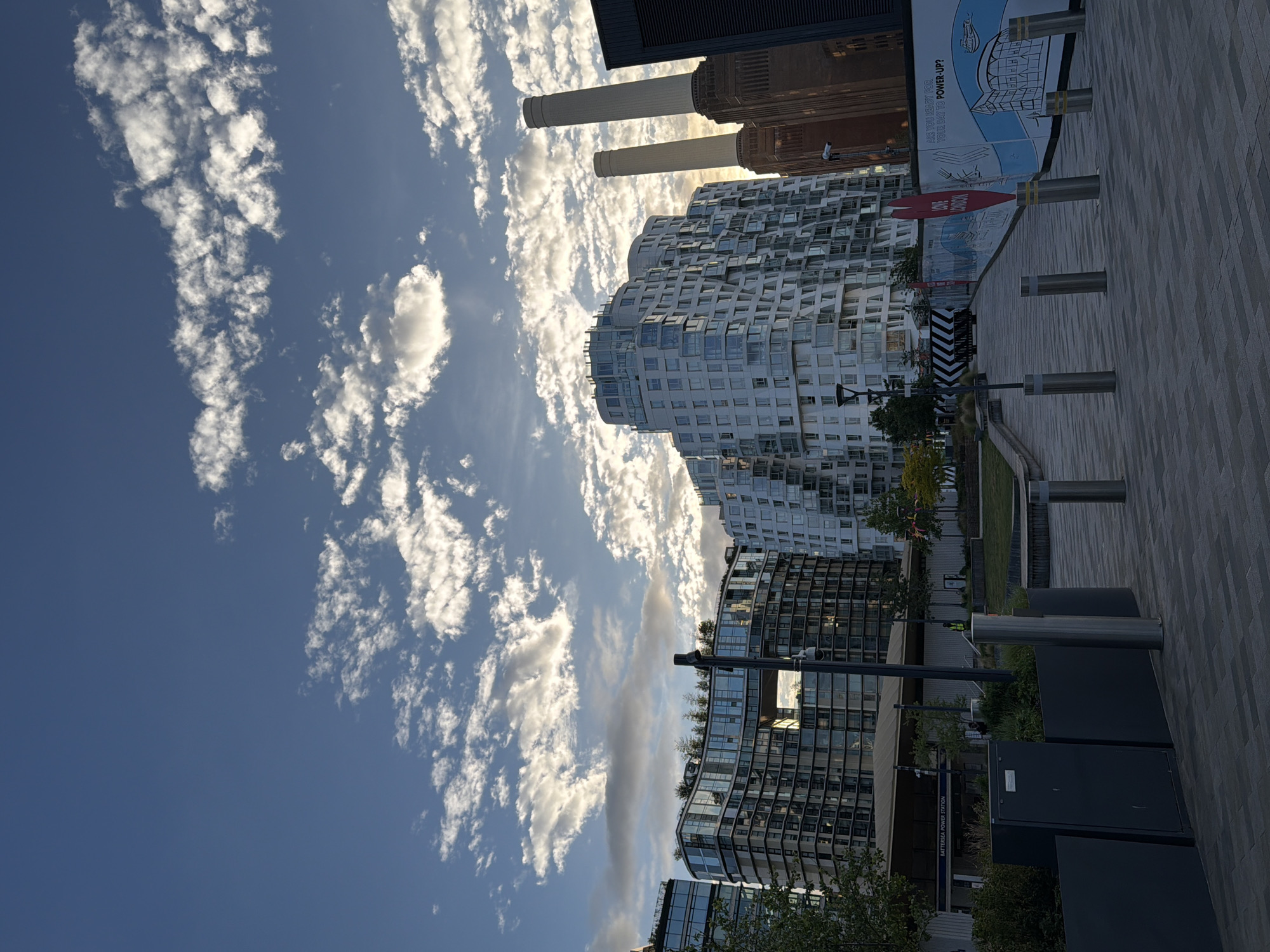 The image depicts the Battersea Power Station in London, United Kingdom, against a blue sky with streaky white clouds. Modern residential buildings with wavy facades are positioned in front of the iconic power station. The foreground features a pedestrian walkway with grey paving stones, bollards, planters with greenery, and public utility boxes. Some signage and infrastructure are also visible along the walkway. The overall atmosphere appears bright and sunny.
