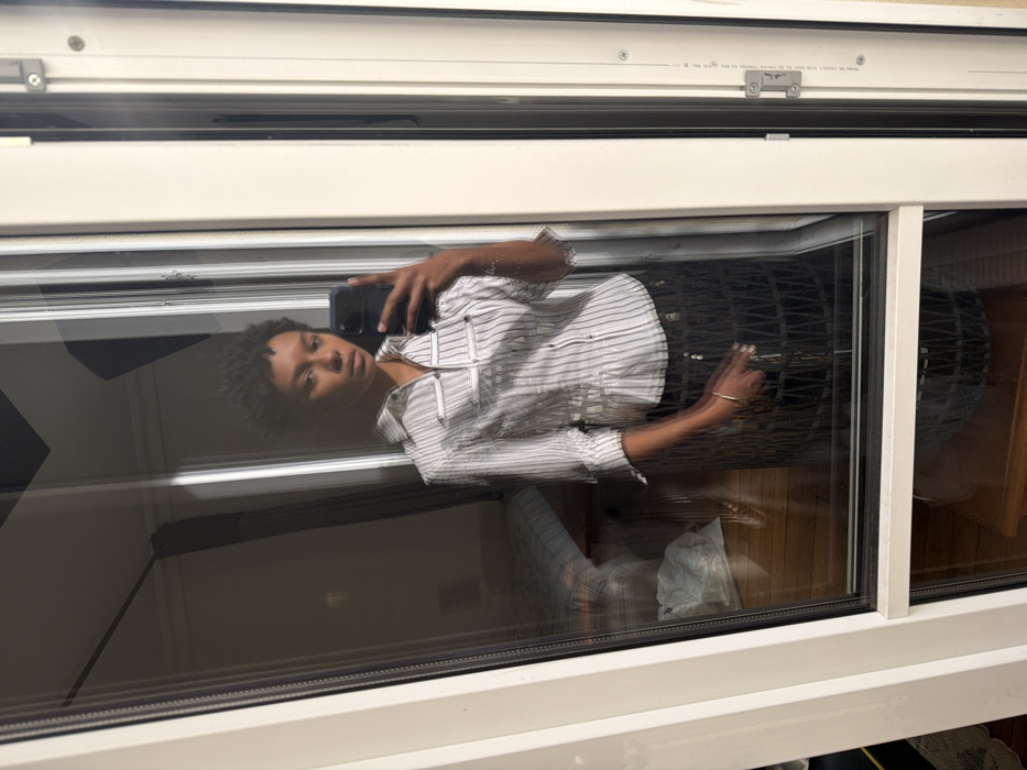 The scene depicts an indoor mirror selfie captured in an interior room located in Paris, France. A young adult female with dark skin and short, dark curly hair stands centrally, facing the camera with a neutral expression. She wears a white and grey vertically striped, short-sleeved collared shirt featuring four horizontal strap details with metallic clasps on the chest and a central closure. A dark, textured, possibly woven or mesh, skirt is visible below her waist. A slender gold-colored bracelet adorns her left wrist. She holds a dark-colored smartphone in her right hand, extended to capture the self-portrait reflection. The reflection is cast upon a large, clear glass pane, likely part of a white-framed French window or balcony door, with a white frame and visible silver hardware elements on the right edge. The reflected background reveals a room with light-colored walls and wooden flooring. A bed with a light-colored striped duvet is partially visible on the left side of the reflection. Overhead architectural details or light fixtures are also reflected.