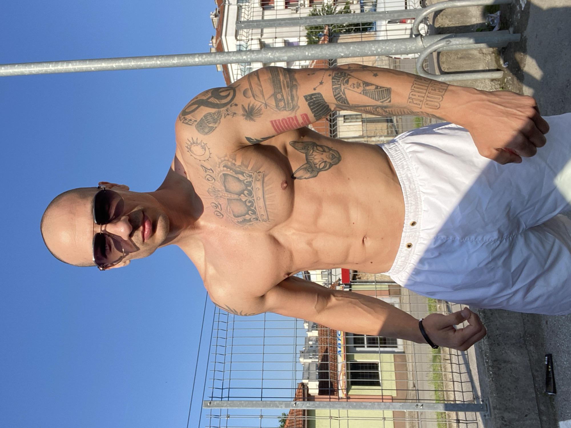 A muscular, bald man is captured in a sunlit outdoor setting, likely in Pleven, Bulgaria. He is seen from the waist up, shirtless, and wearing white shorts and dark aviator-style sunglasses. The man stands with a confident, intense expression, looking directly at the camera. His physique is well-defined, with visible abdominal muscles and prominent veins on his arms.
His body is extensively tattooed. On his left arm, visible designs include a microphone, a palm tree, and the word "WORLD" in block letters (appears red). His left chest features a large, ornate crown or frame tattoo, and near his left shoulder, there is a sun and crescent moon design. On his right pectoral, a distinctive cat-like or mythological creature is tattooed. His right forearm clearly displays the text "KNOW OVER".
The background suggests an urban environment. A metal fence or barrier runs horizontally across the mid-ground, behind the man. Beyond the fence, there are multi-story buildings, some with visible windows and balconies, and hints of greenery. The sky is clear and bright blue, indicating sunny weather during the daytime. A metal railing is visible in the foreground. The man's right hand, adorned with a black wristband, is relaxed, while his left hand is clenched, suggesting a pose.