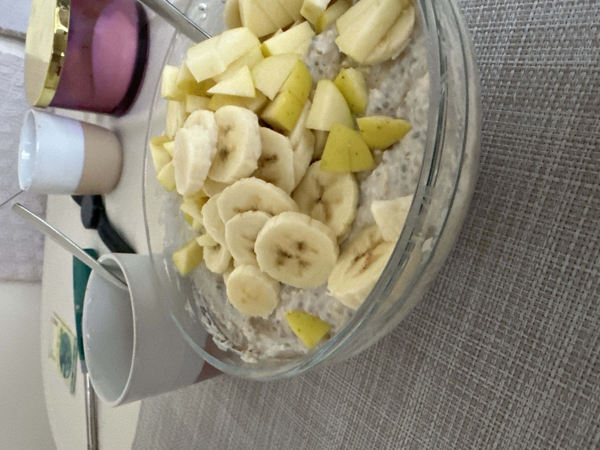 An overhead close-up shot shows a bowl of oatmeal topped with sliced bananas and diced apples, placed on a textured gray placemat. To the left of the bowl, a white and pink mug contains a spoon. In the background, two other cups are visible, one white and tan, the other a pink candle in a glass container with a gold lid. A metallic object resembling a knife or spoon lies on the table behind the mug. The setting appears to be indoors, possibly a home or cafe, with a hint of currency or paper in the upper left corner. The lighting is natural and bright, suggesting it is daytime. There are no people present in the image. The scene is devoid of any specific Varna landmarks.