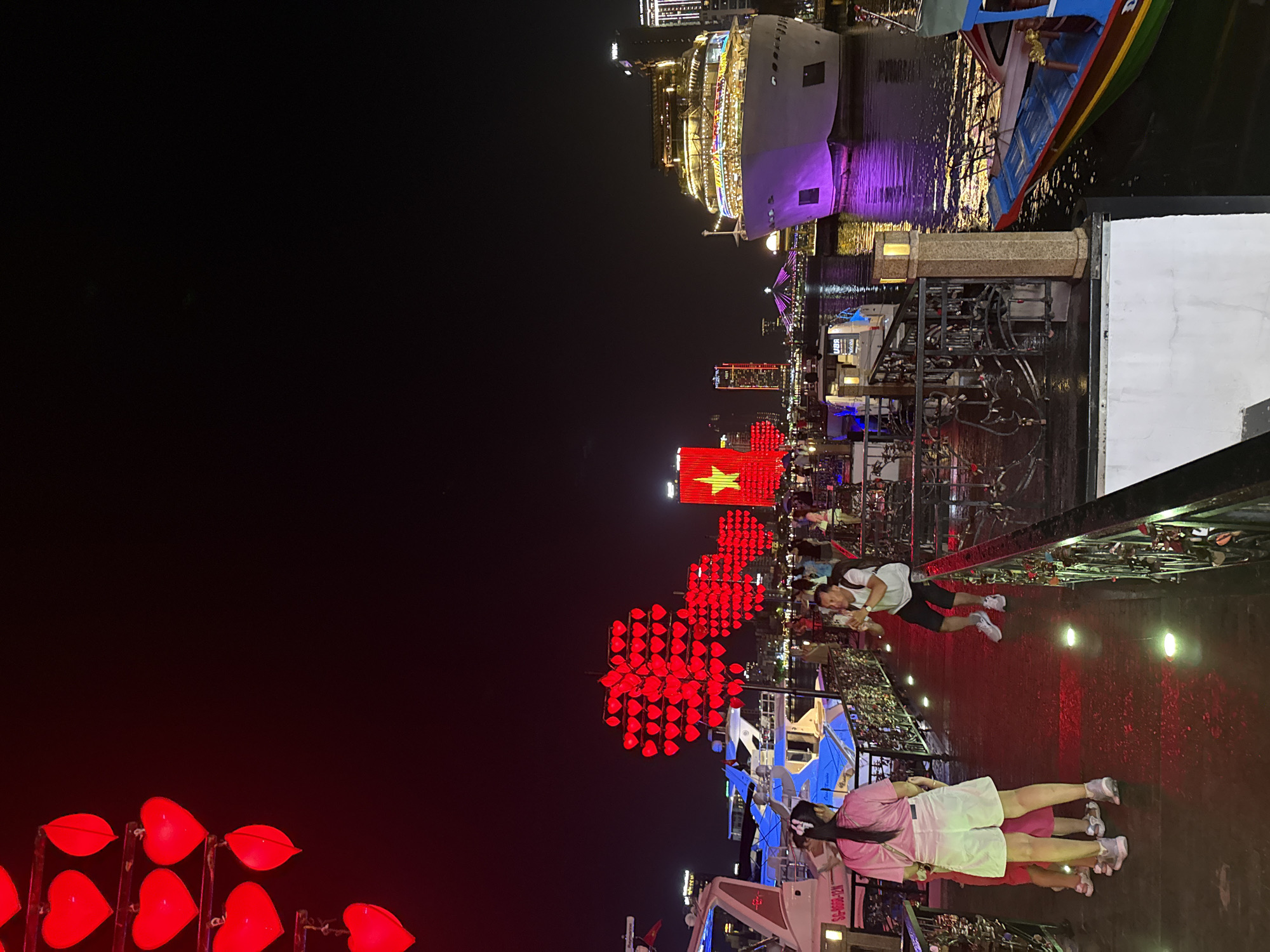 The image depicts a nighttime scene along the Hàn River in Đà Nẵng, Vietnam. Red heart-shaped lanterns and the Vietnamese flag illuminated in red and yellow are prominent. Several people are visible, including two women in pink and white attire in the foreground and a man in a white shirt and dark shorts walking along the walkway. The Hàn River Bridge and a large cruise ship with illuminated decks are visible in the background. Boats are docked along the river. The environment is a combination of urban landscape and waterfront.