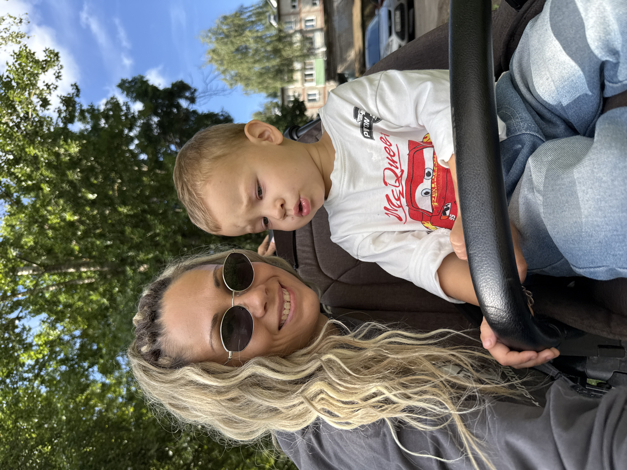A medium shot captures a woman and a young child in a stroller outdoors, possibly in the city of Rusanovo, Russia. The woman, with blonde dreadlocks and sunglasses, smiles at the camera. The child, wearing a white long-sleeved shirt with a "McQueen" graphic, looks to the side with a slightly pursed mouth. They are seated in a stroller with a black handle in the foreground. Behind them, green trees and a blue sky with clouds are visible, with buildings and a parked car in the background. The scene suggests a casual outdoor outing on a sunny day.