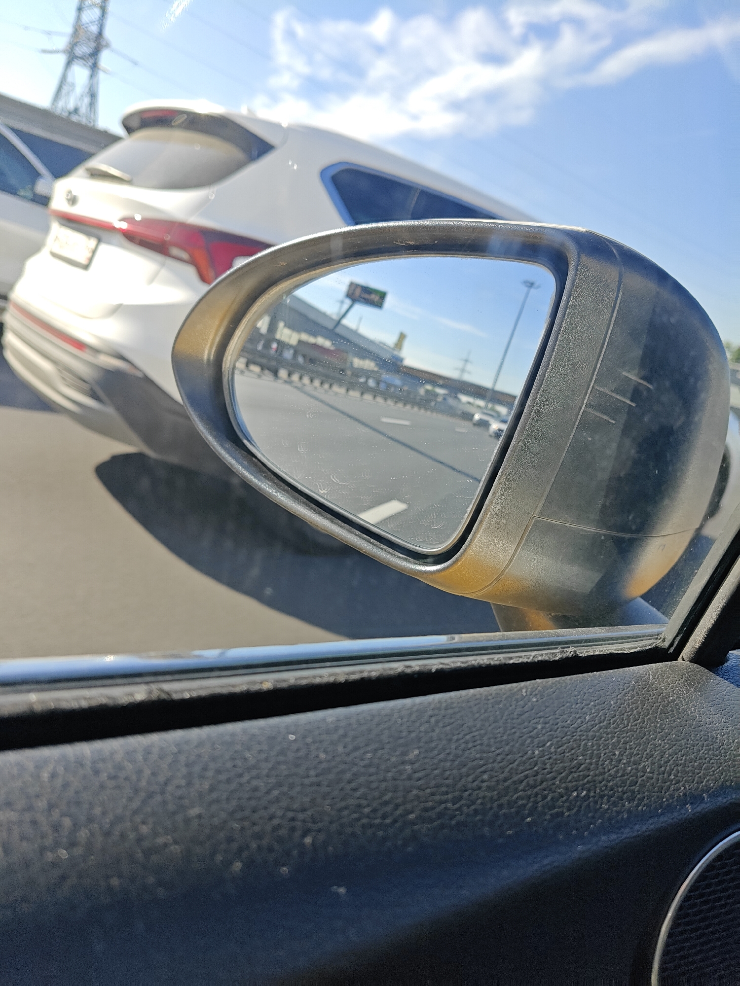 The image is a first-person perspective from inside a car, looking at the side mirror. The car is in motion on a highway, as indicated by the road markings and other vehicles visible in the reflection. The time of day is daytime, with a clear blue sky and scattered clouds.
In the foreground, the textured black interior of the car's door panel is visible. The side mirror itself is a dark gray color with a metallic gold trim around the reflective surface. The mirror's surface is slightly dirty, with visible smudges and water spots.
In the reflection of the side mirror, the highway stretches out ahead. Several cars are visible, including a white SUV that appears to be in the adjacent lane. The SUV has a prominent spoiler and red taillights. In the distance, there are some buildings and industrial structures, suggesting an urban or suburban environment. The presence of a large electrical pylon on the left side of the frame further points to an infrastructure-heavy area.
The location is Mytishchi, Russia. The specific context is likely a typical commute or travel on a highway within the city or its outskirts, given the road conditions, traffic, and visible infrastructure. No individuals are clearly identifiable in the image. The overall scene depicts a moment of travel, capturing the mundane but informative view from a car's side mirror.