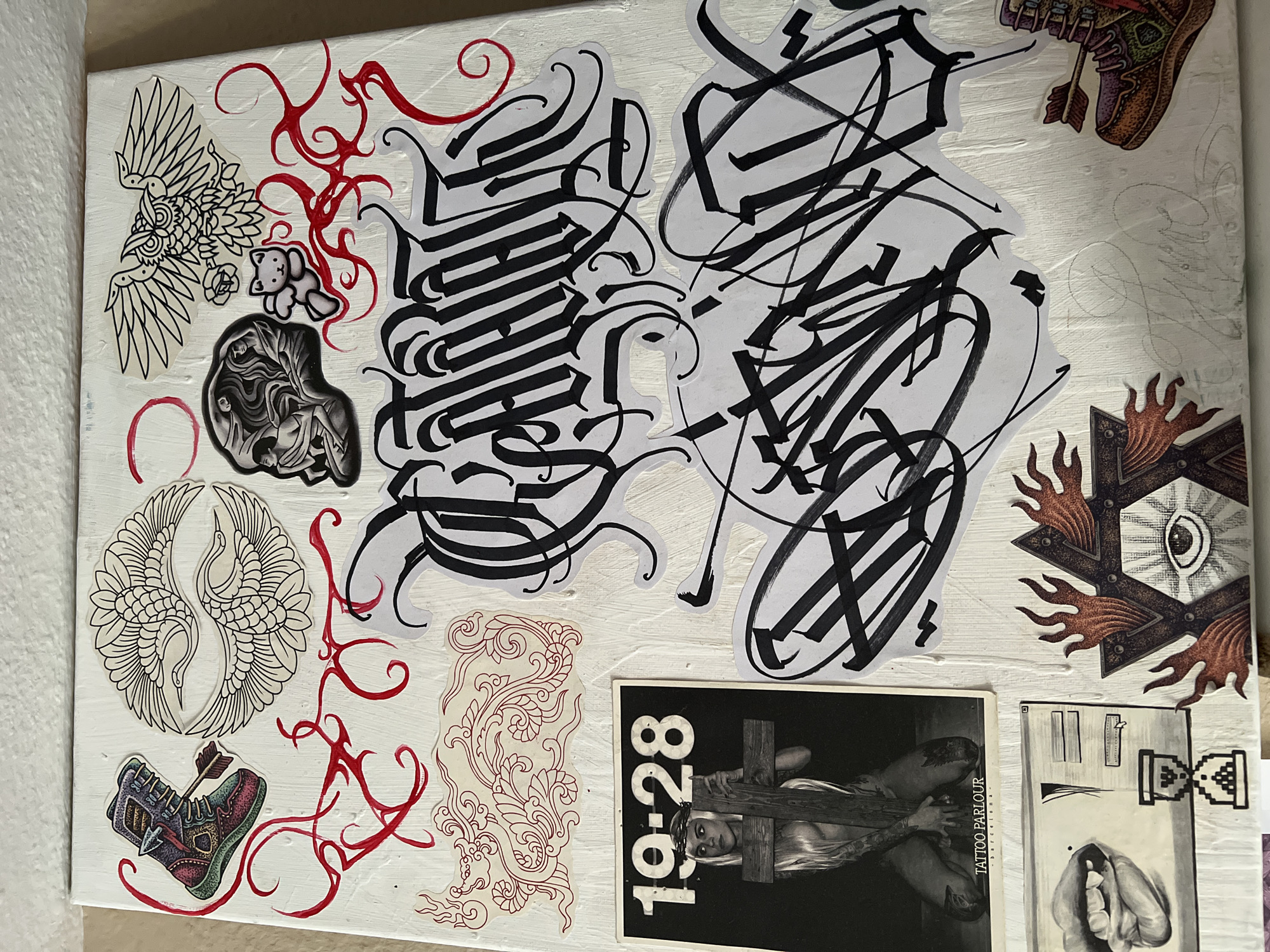 The image shows a collage of images, predominantly black and white, on a white background.
The collage consists of illustrations and photographs, including:
* Two large black calligraphy designs.
* Illustrations of winged creatures, a teddy bear, a boot with an arrow, a floral pattern, and a shield with an eye.
* A black and white photograph with "19-28 TATTOO PARLOUR Barcelona" written on it.
* A drawing of an hourglass.
* An image of two cranes with their wings outstretched.
* Red embellishments that look like doodles or artistic flairs throughout.
There is no information about the location being Madrid, Spain.