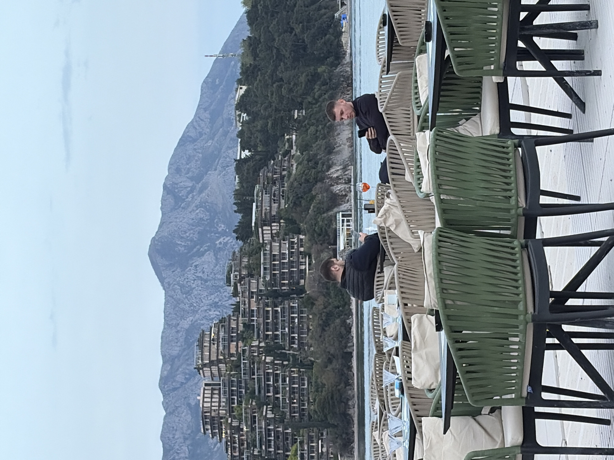 The scene shows a row of outdoor chairs on a terrace, with two men in dark clothing seated. One man is looking at his phone. In the background, there are multi-story buildings on a hillside and a large mountain. The image is likely taken in or near Budva, Montenegro, given the architecture and mountainous terrain. There is also an aperol spritz drink on the table.