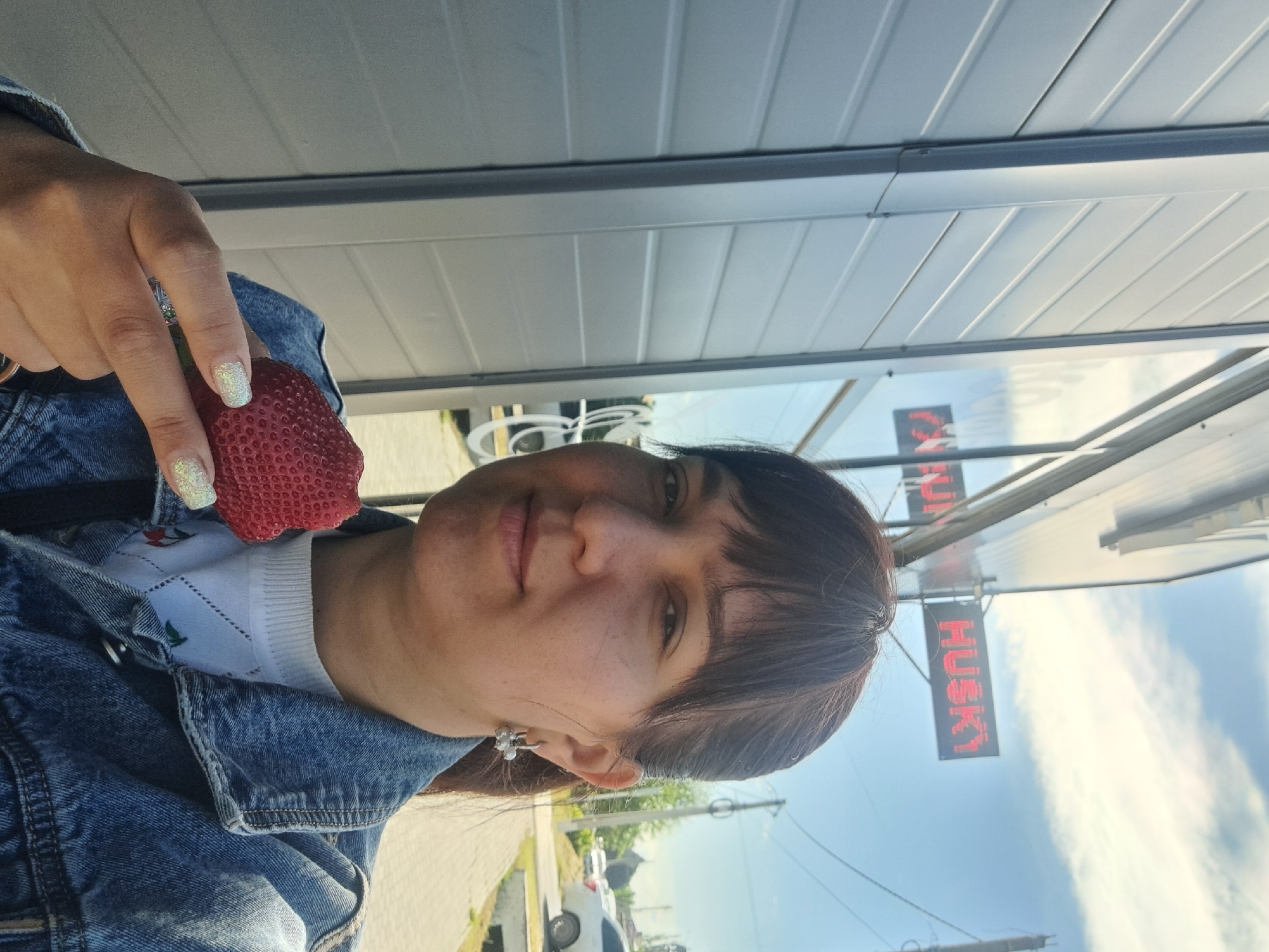 A woman is holding a red strawberry. She is standing outside near a building with a sign that says "HUSKY" in red letters. The location is Leningradskaya, Russia.