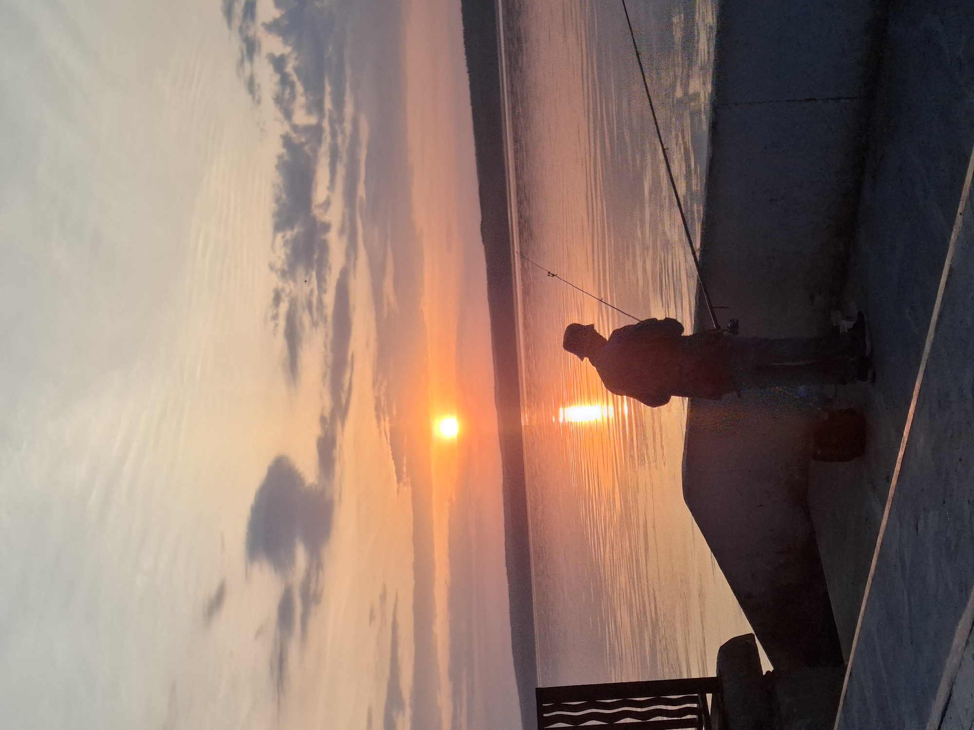 A horizontal shot depicts a person fishing at sunset on a body of water. The location is identified as Chaikovsky, Russia. The person is on a concrete structure, possibly a pier or embankment, with fishing rods extending over the water. The sky is filled with clouds colored by the sunset, reflected in the water. A dark, partially obscured object with a wavy design is visible near the bottom of the frame.