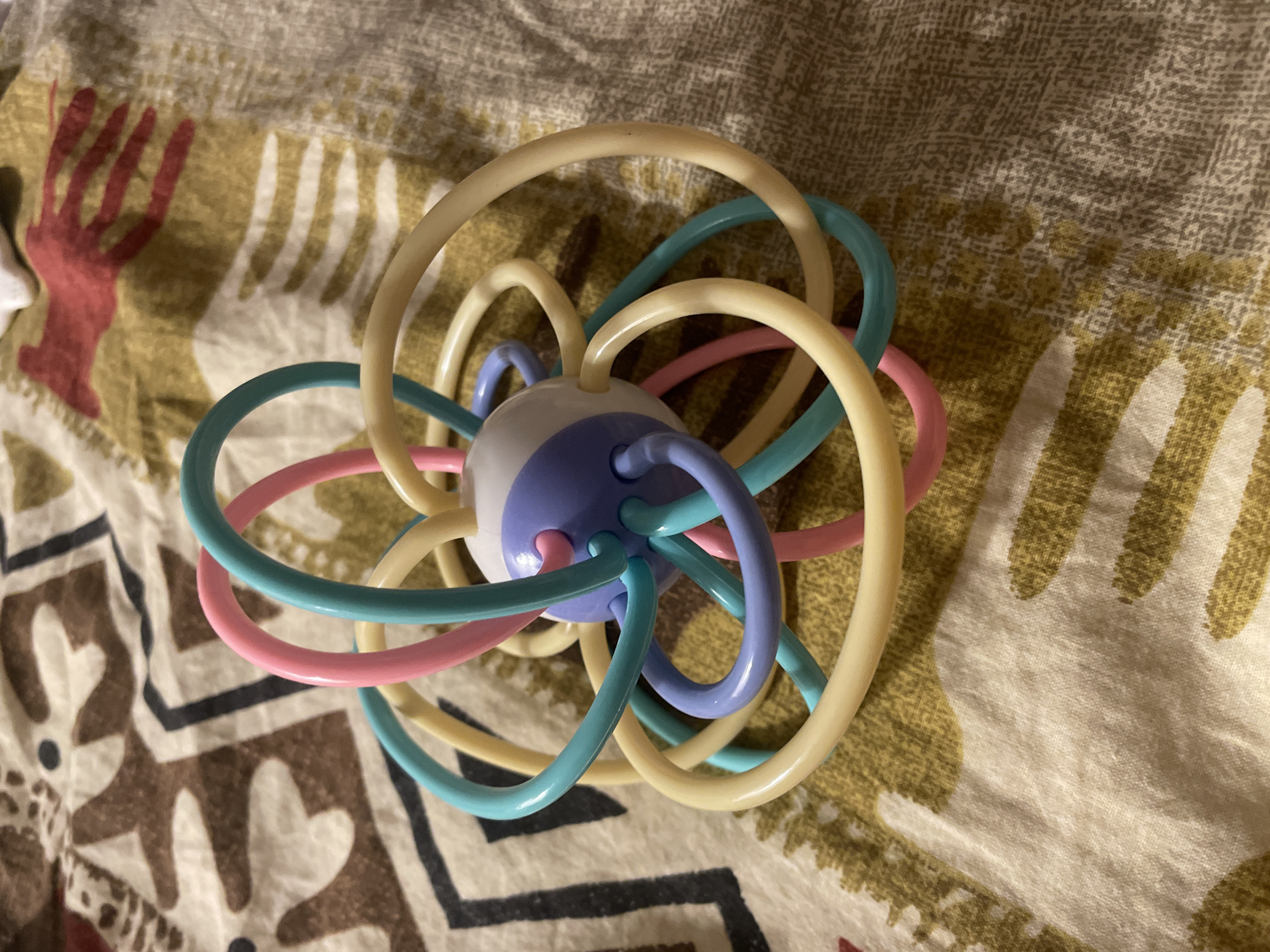 The image shows a close-up, high-angle shot of a colorful baby rattle toy on a patterned fabric surface. The rattle is spherical with flexible plastic loops in various pastel colors: yellow, pink, blue, and lavender. The loops are attached to a central, multi-colored sphere, which appears to be made of plastic. The fabric beneath the toy has a complex, somewhat abstract pattern in shades of brown, beige, and white, with some red accents. The lighting suggests an indoor setting. There is no indication of a specific location in Zemun Urban Municipality, Serbia, in the image itself.