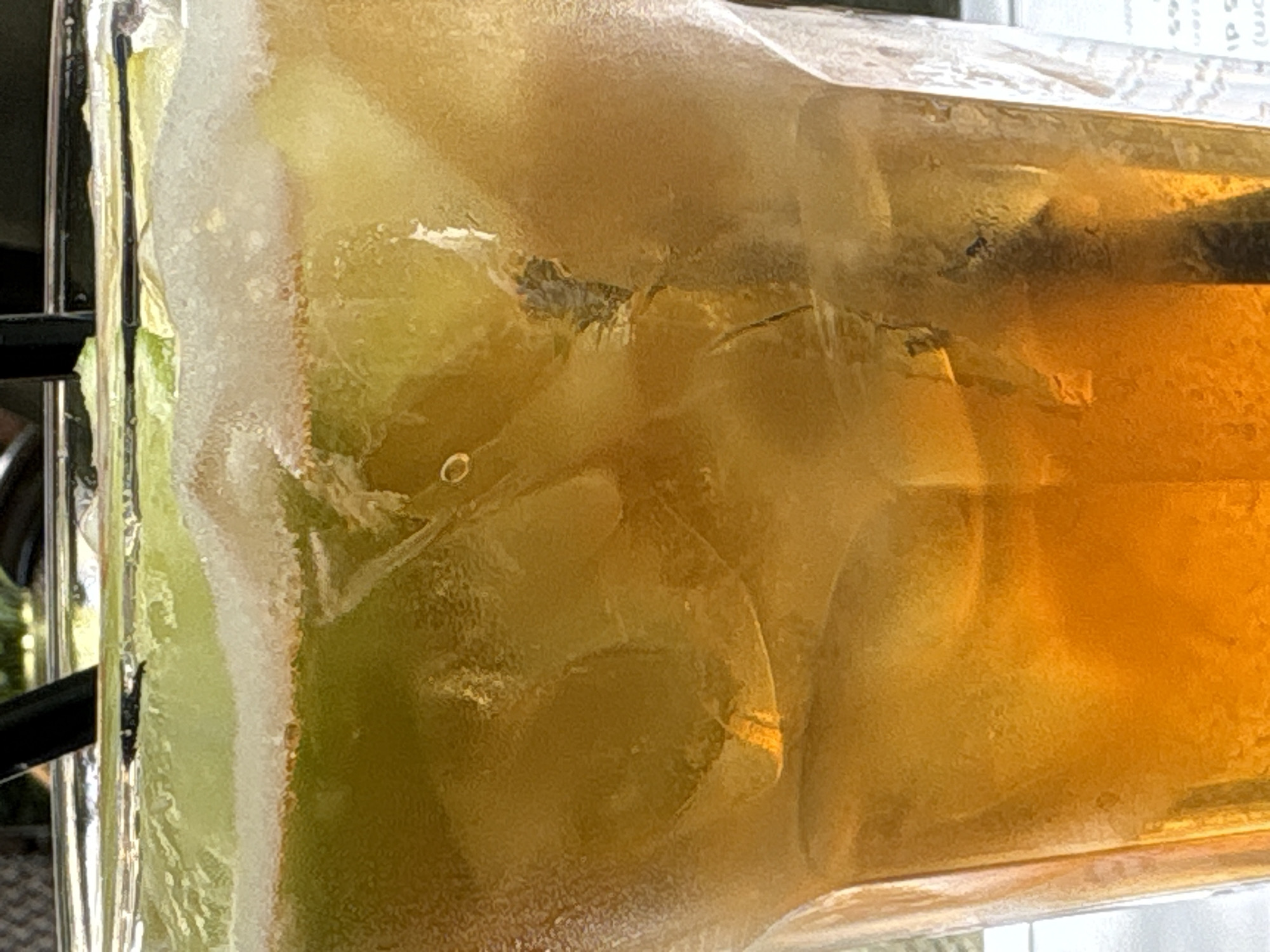 The image is a close-up of a tall glass filled with an amber-colored beverage and ice. There are green slices of fruit floating on top, along with a layer of foam. Two black straws are visible protruding from the top of the glass. The glass itself appears to be sweating, indicating a cold drink. The background is blurred, but suggests an outdoor setting with greenery. There is no discernible location in the city of Belgrade, Serbia, visible in this image.