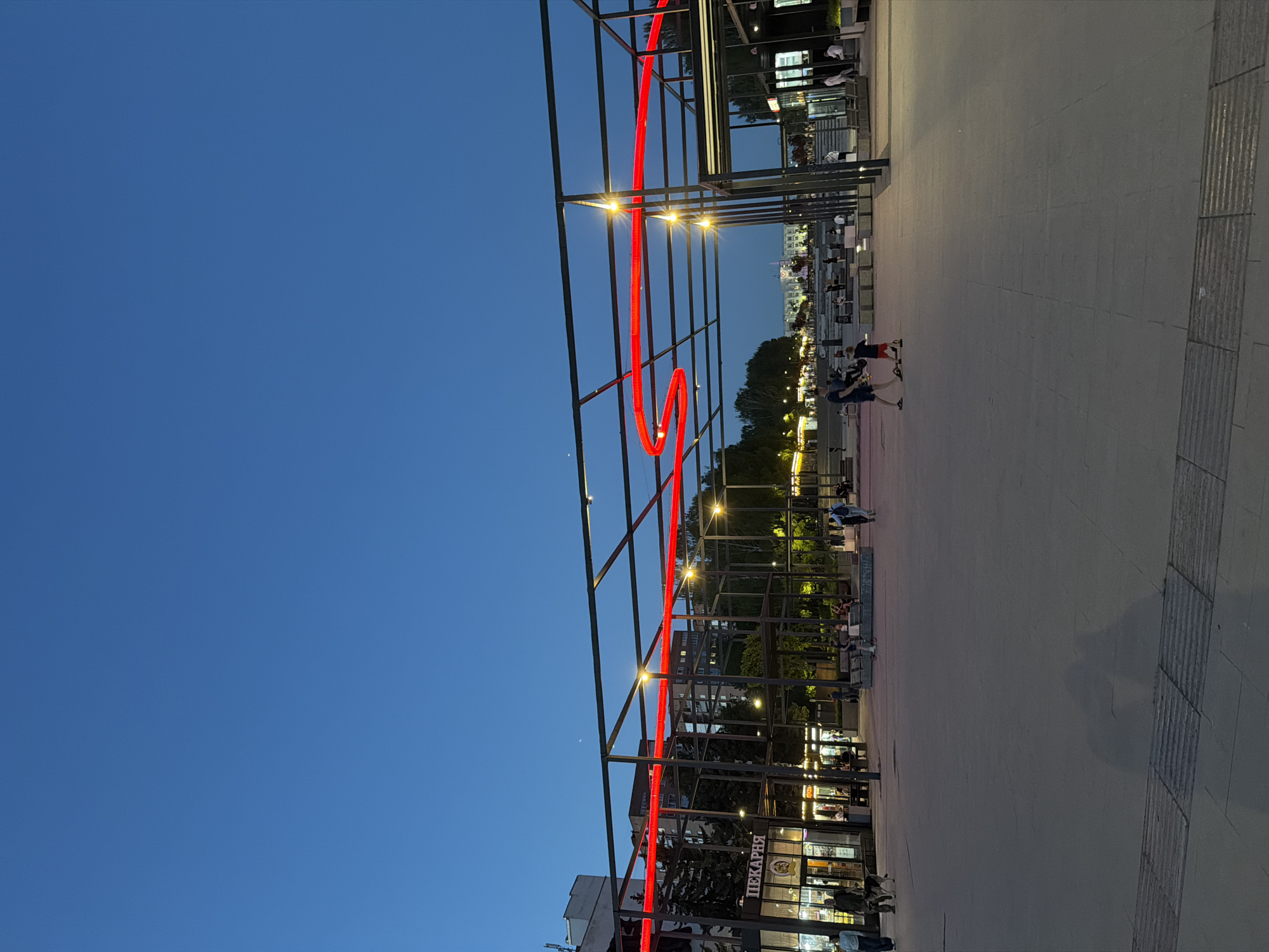 The image shows a public square in Izhevsk, Russia, at dusk. A metal framework, illuminated by a red neon light forming a stylized “S” shape, stands prominently in the foreground. The framework also has built-in lighting. The background includes trees and city buildings with illuminated windows. People are walking and standing in the square. Shops with signage in Russian ("ПЕКАРНЯ") are visible. The pavement is made of light-colored slabs. The sky is a deep blue, transitioning to lighter shades near the horizon.